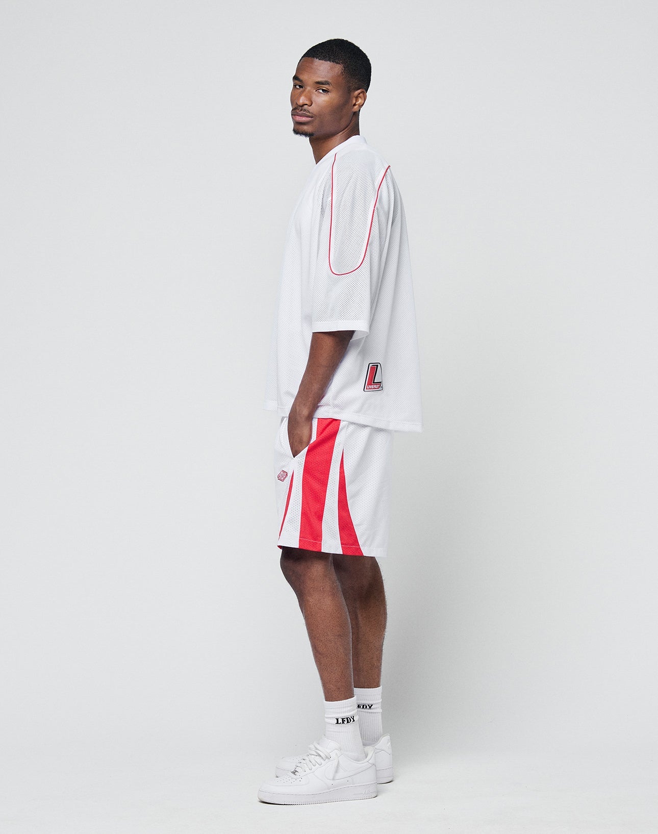 A man with short hair stands sideways against a plain background, wearing the LIVE FAST DIE YOUNG Basic Mesh Jersey in white with red accents, paired with white socks and sneakers, and maintains a neutral expression.