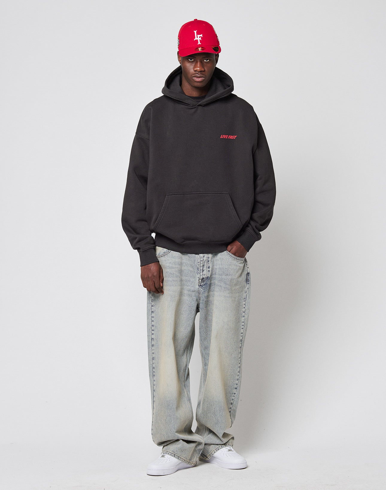 A person stands against a plain backdrop wearing an oversize fit black LFDY Globe Hooded by LIVE FAST DIE YOUNG with front and back print, light blue baggy jeans, white sneakers, and a red baseball cap with white lettering.