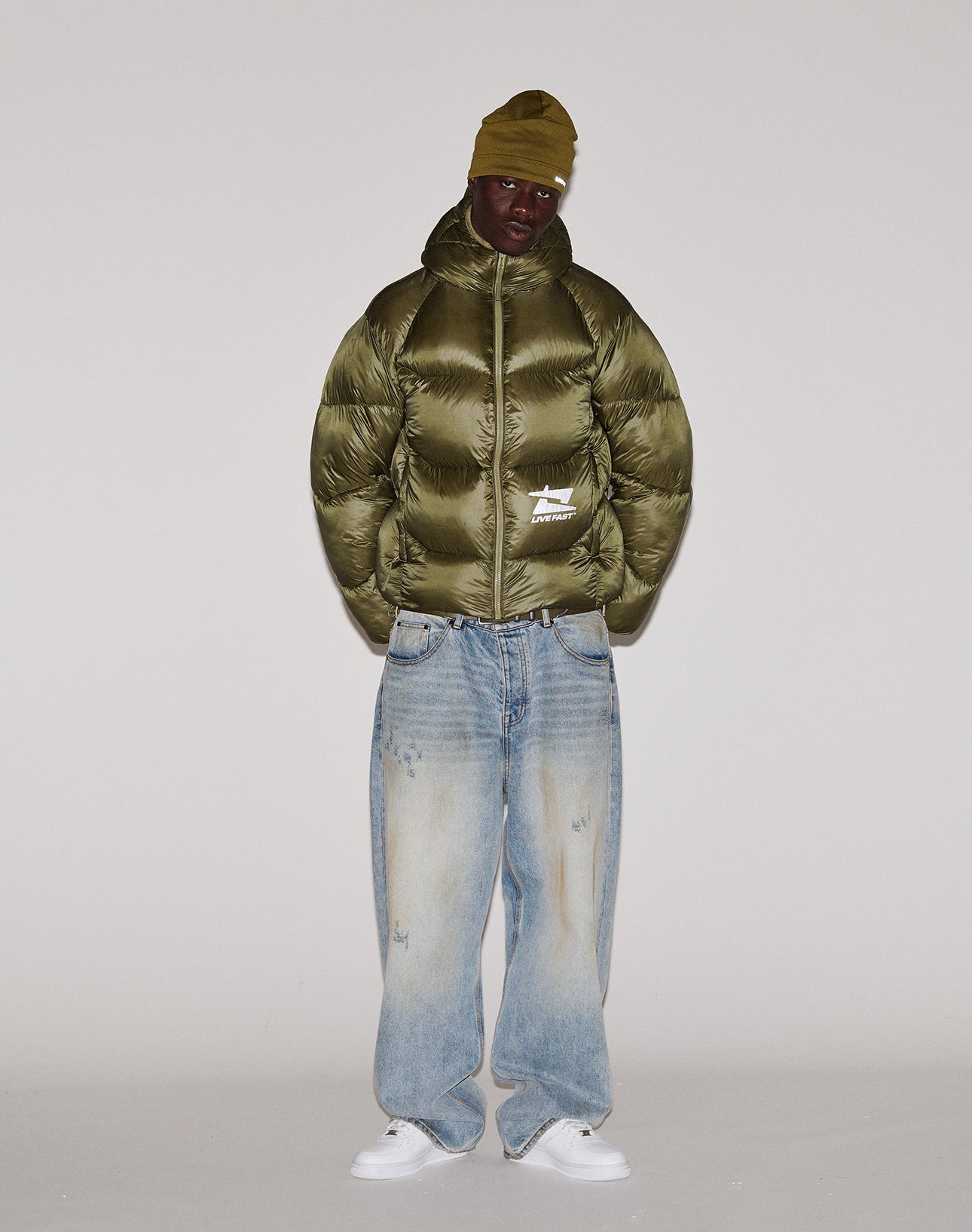 A person wearing a Live Fast Puffer Jacket by LIVE FAST DIE YOUNG, an olive beanie, light blue baggy jeans, and white sneakers stands against a plain, light-colored background.