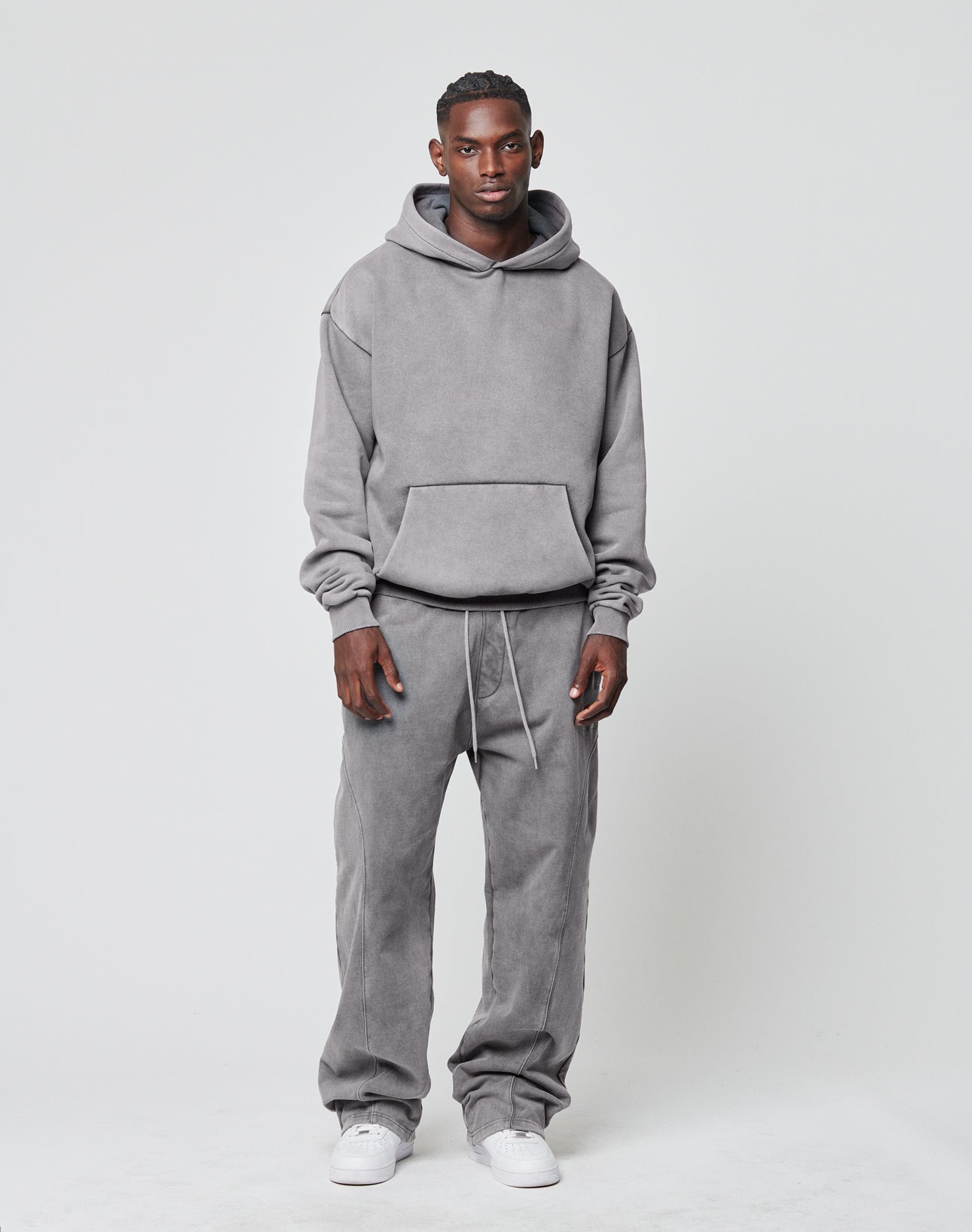 A man stands against a light background, facing forward with a neutral expression. He wears the LIVE FAST DIE YOUNG Basic Blank Hooded in gray, matching gray drawstring sweatpants, and white sneakers.