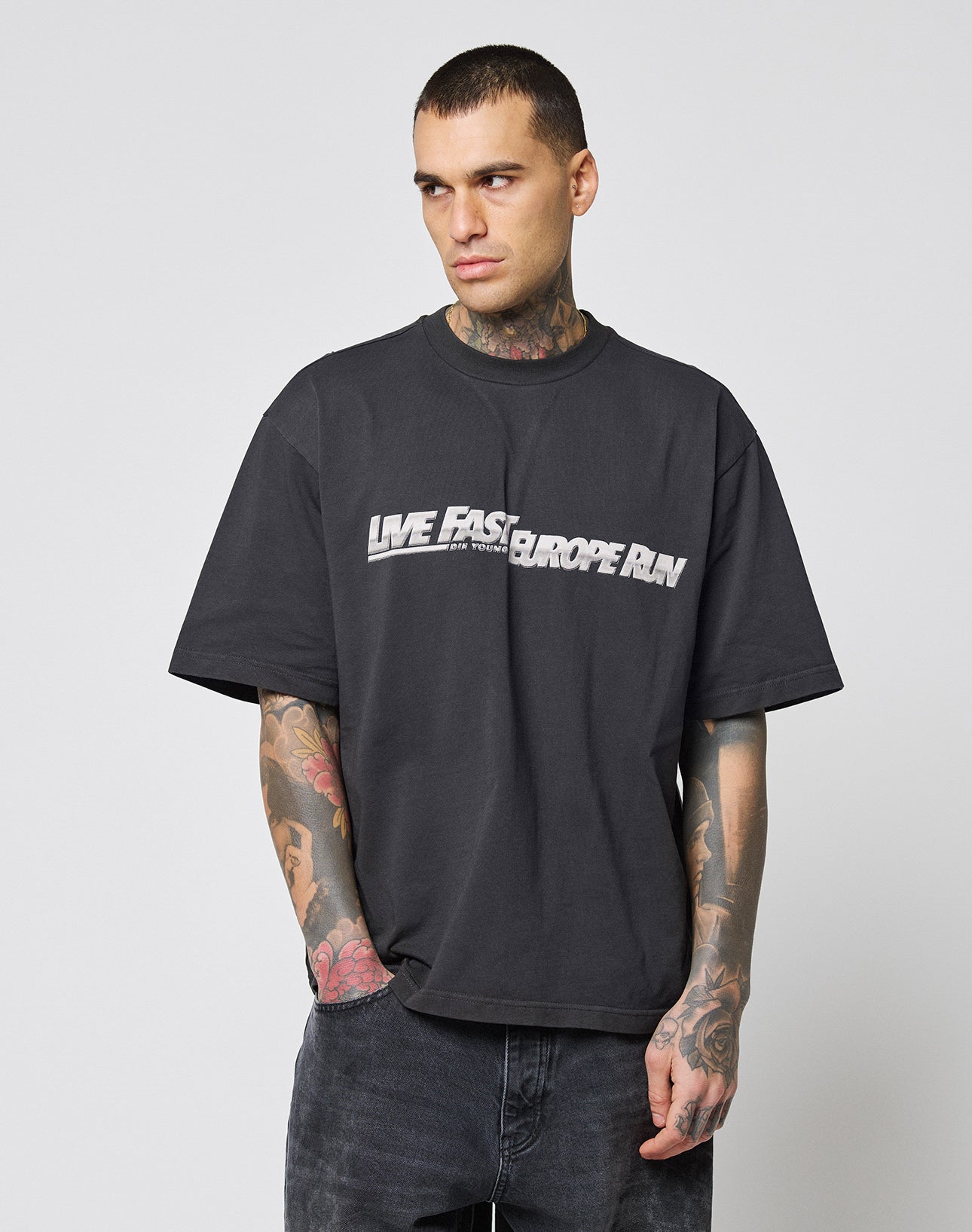A tattooed person stands with hands in pockets, looking sideways against a plain background, wearing the LIVE FAST DIE YOUNG LFDY Europe Run Tee—an oversized black cotton T-shirt featuring “LIVE FAST EUROPE RUN” in white text.