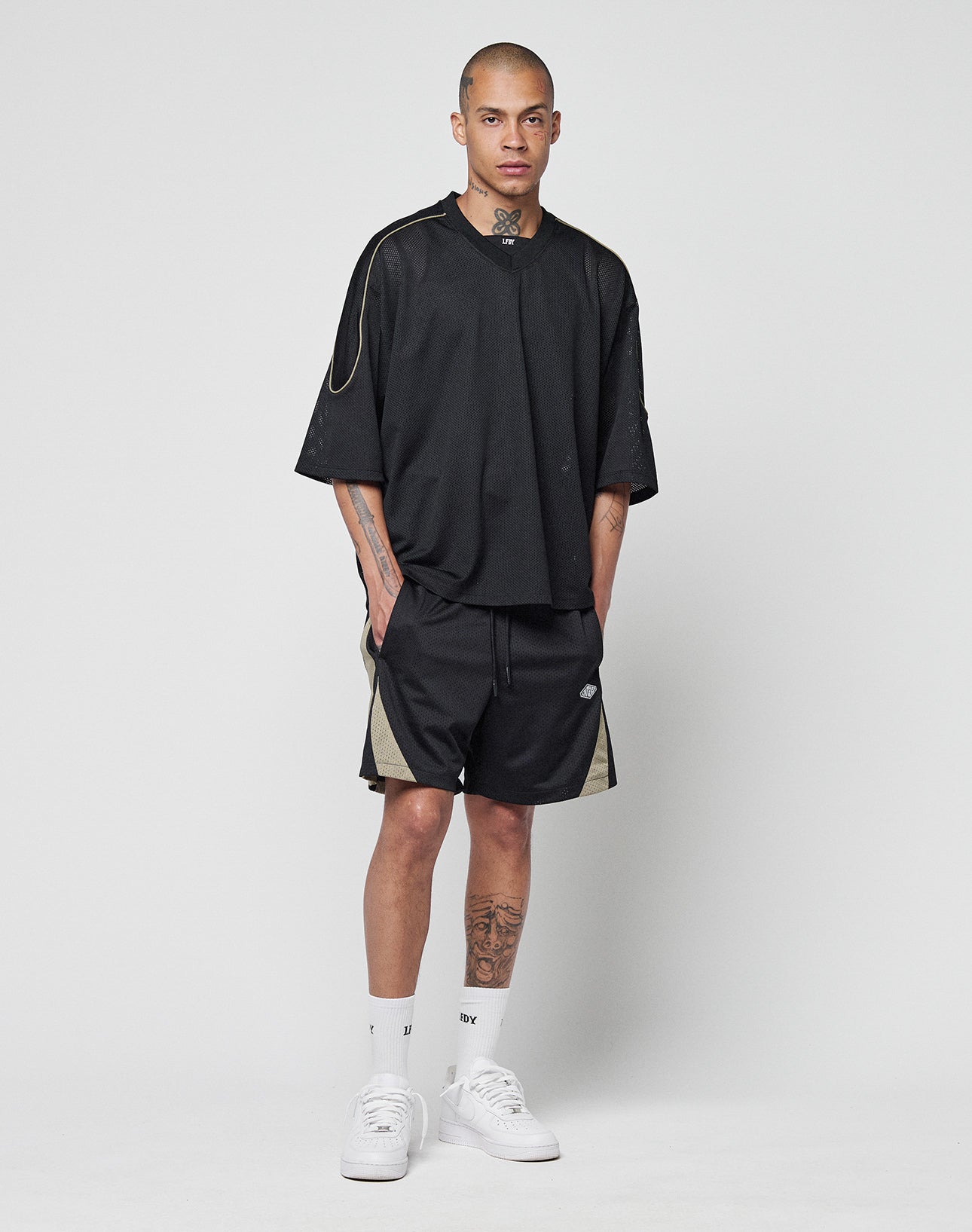 A man with tattoos stands against a plain white background wearing a black LIVE FAST DIE YOUNG Basic Mesh Jersey, black shorts with beige contrast piping, white crew socks, and white sneakers. He has his hands in his pockets and a neutral expression.