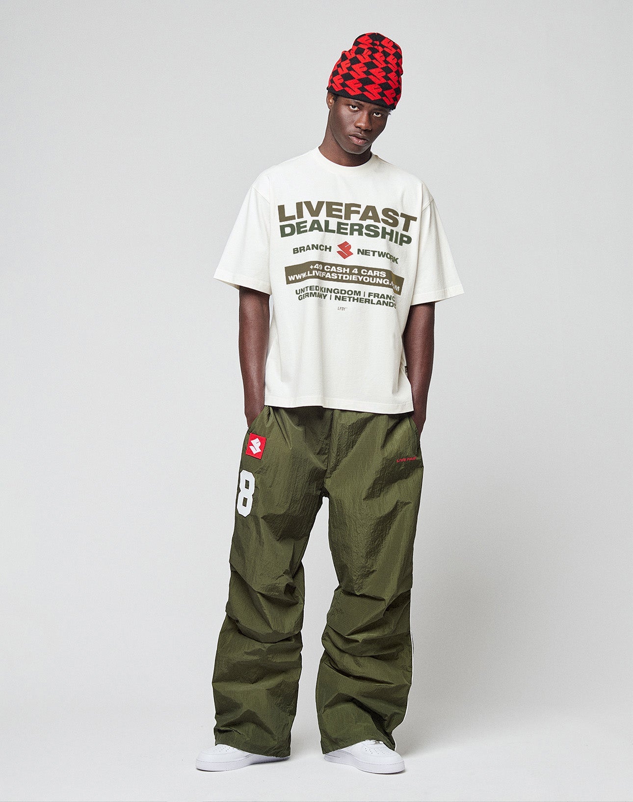 A man in a red and black checkered beanie, an oversized white LIVE FAST DIE YOUNG logo tee, LFDY Nylon Trackpants in olive green with 88 and patches, and white sneakers stands against a plain light gray background.