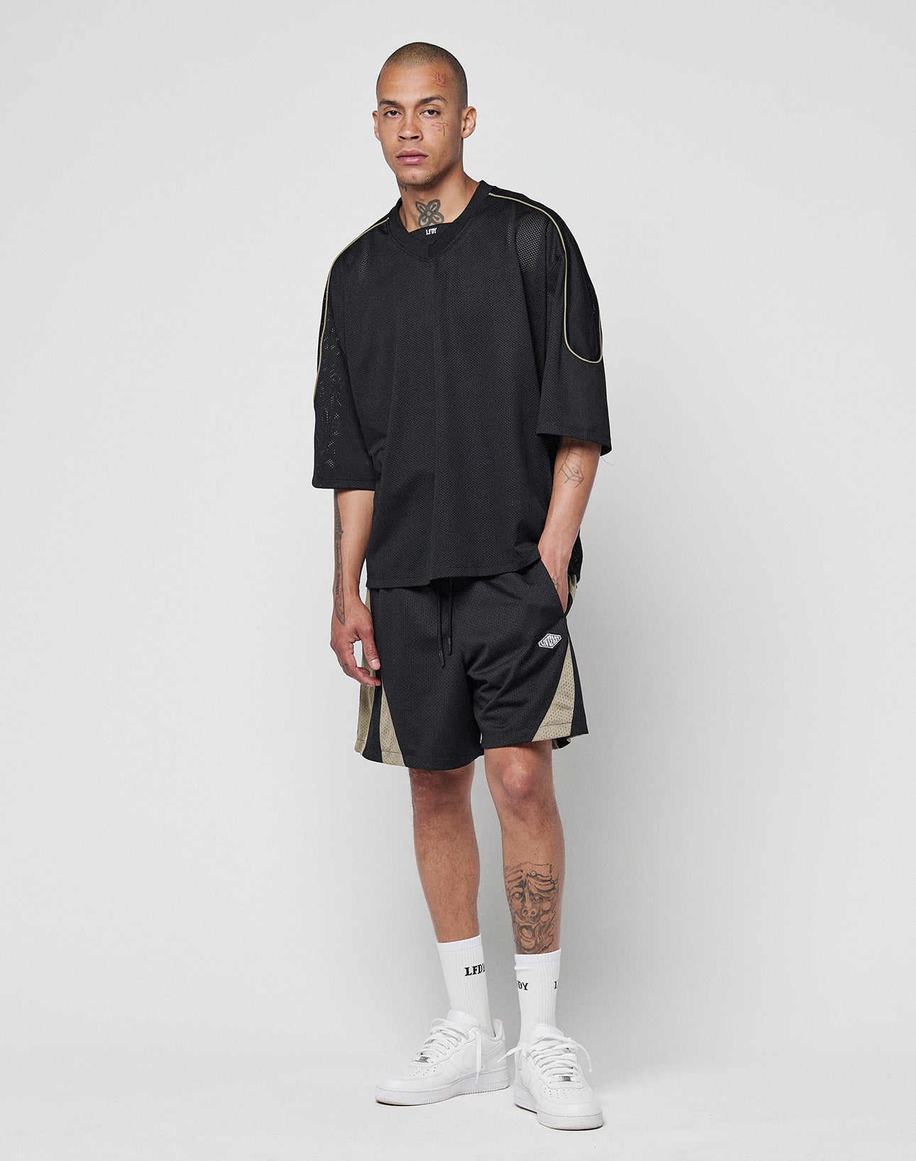 A man with short hair and visible tattoos on his neck and leg wears a loose black T-shirt with a logo patch, LIVE FAST DIE YOUNG Striped Mesh Shorts, white crew socks, and white sneakers against a plain white background.