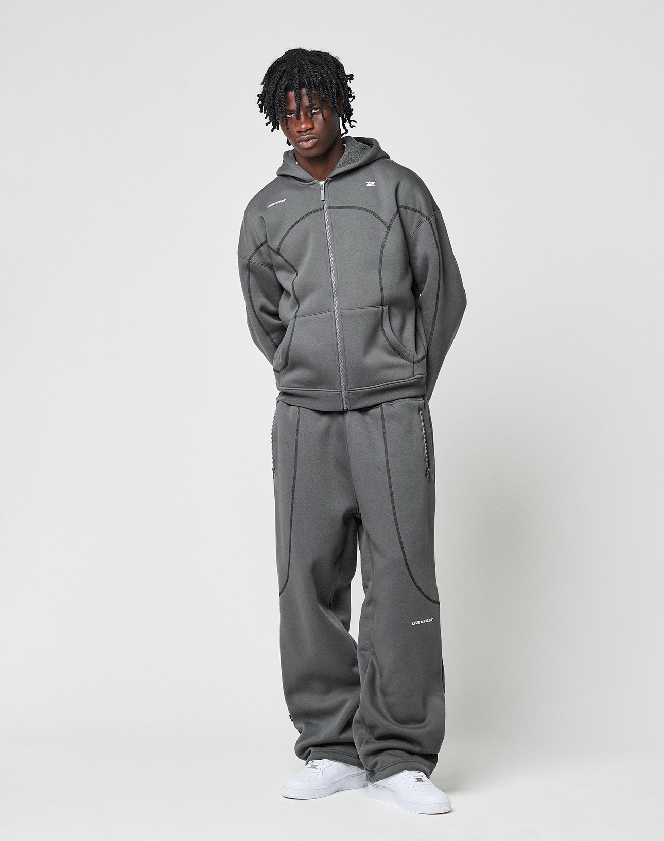 A person with short, curly hair stands against a white background, wearing LIVE FAST DIE YOUNGs LFDY Active Fleece Jogger in a wide fit, paired with a grey hooded zip-up and white sneakers. Their hands are behind their back.