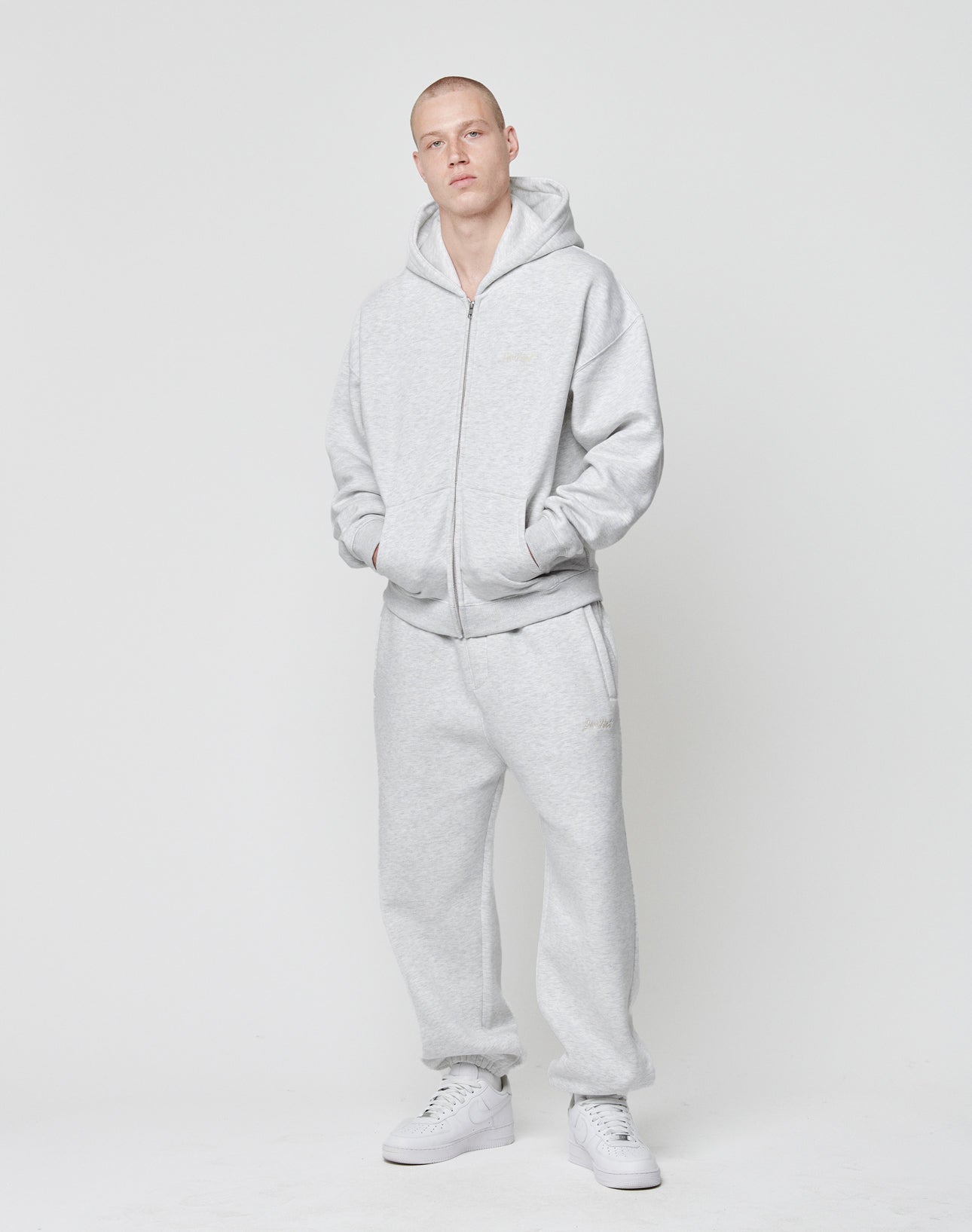 A person stands against a white background, wearing the LIVE FAST DIE YOUNG Basic Script Hooded Zip and matching sweatpants in light gray with subtle logo embroidery, made from a soft cotton-polyester blend. Their hands are in the hoodie pockets.