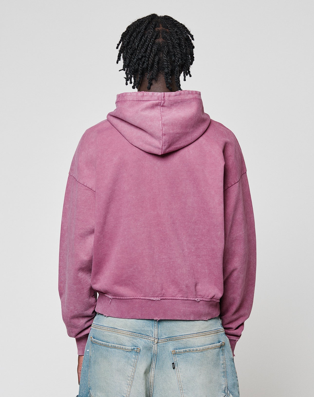 A person with short, twisted hair is seen from behind wearing the LIVE FAST DIE YOUNG Live Fast Rivet Hooded Zip in faded mauve (Oversize Fit), paired with light blue jeans, standing against a plain light gray background.