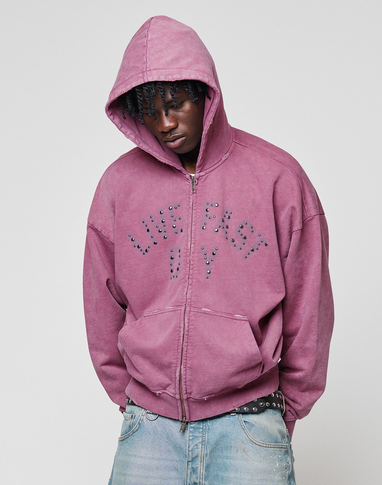 Wearing the LIVE FAST DIE YOUNG Live Fast Rivet Hooded Zip in purple, styled with light blue jeans and a studded belt, a person stands with head slightly bowed against a plain background.