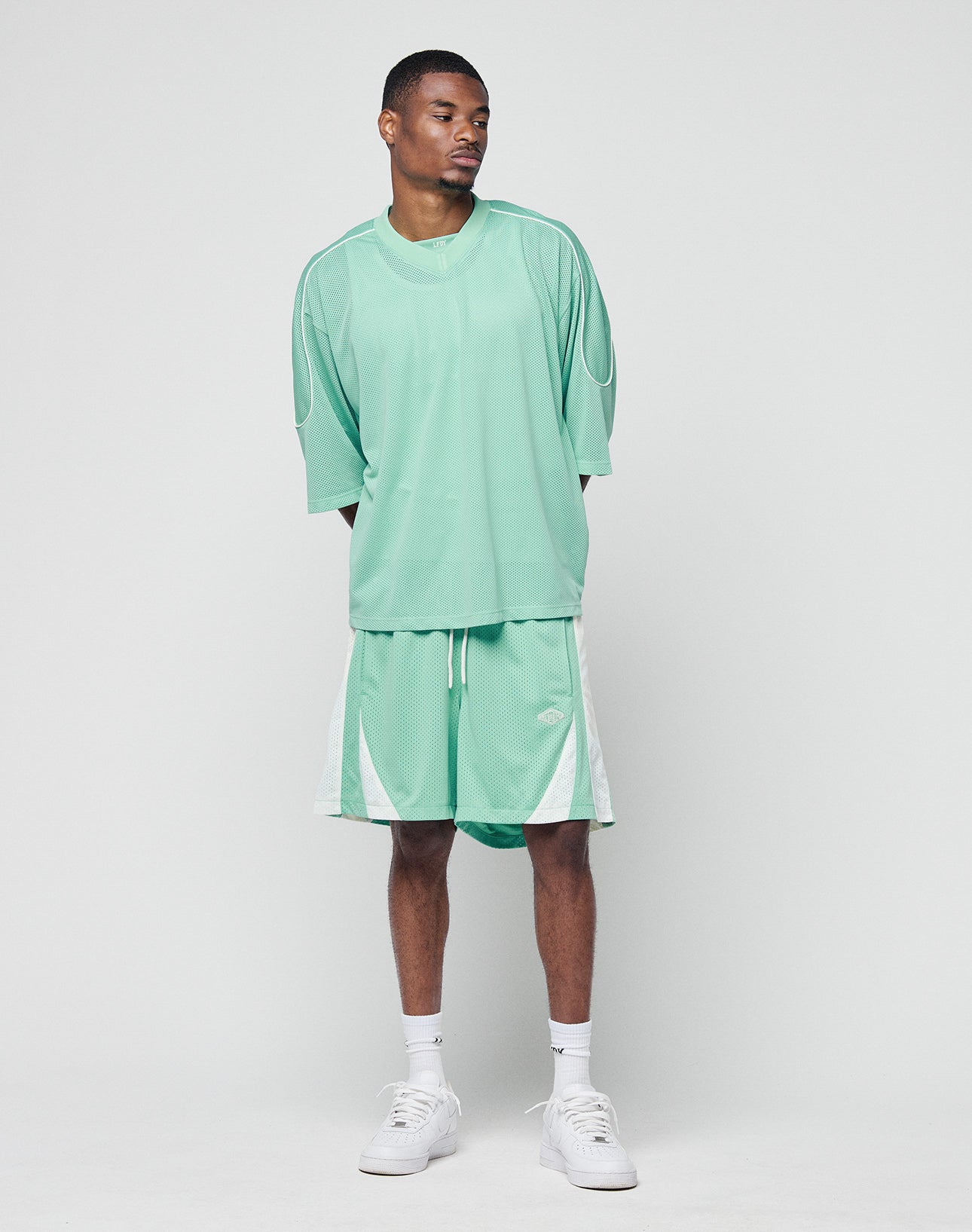 A man stands against a light gray background, wearing an oversize mint green Basic Mesh Jersey and matching shorts with white accents from LIVE FAST DIE YOUNG, paired with white socks and sneakers. He looks slightly left with a neutral expression.