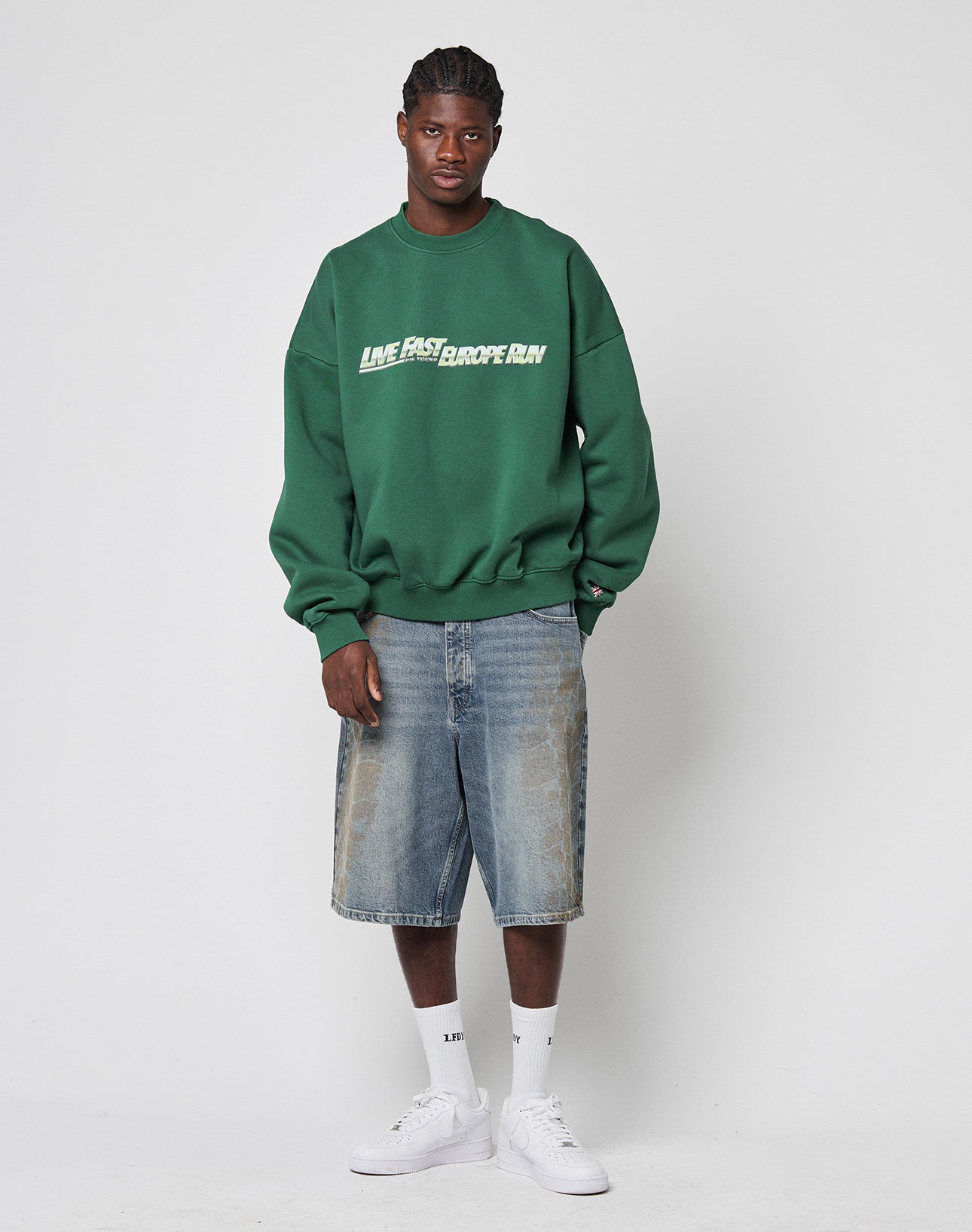 A man with braided hair and a neutral expression stands against a plain background, wearing a green 100% Baumwolle sweatshirt, LIVE FAST DIE YOUNG LFDY Tire Tracks Jorts, white crew socks, and white sneakers.