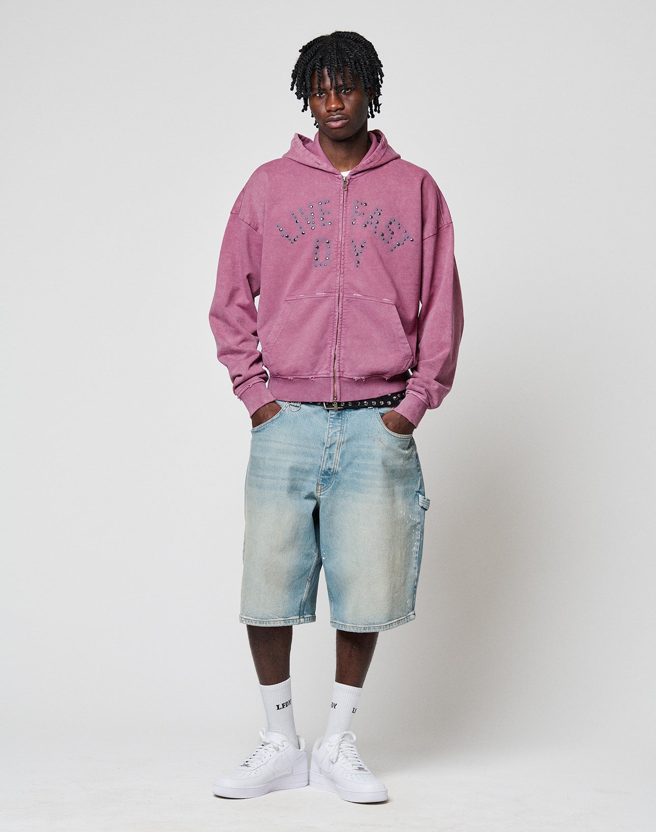 A person stands against a plain background wearing a pink LIVE FAST DIE YOUNG Live Fast Rivet Hooded Zip with handgefertigtem Sprayeffekt, light blue baggy denim shorts, white crew socks, and white sneakers.