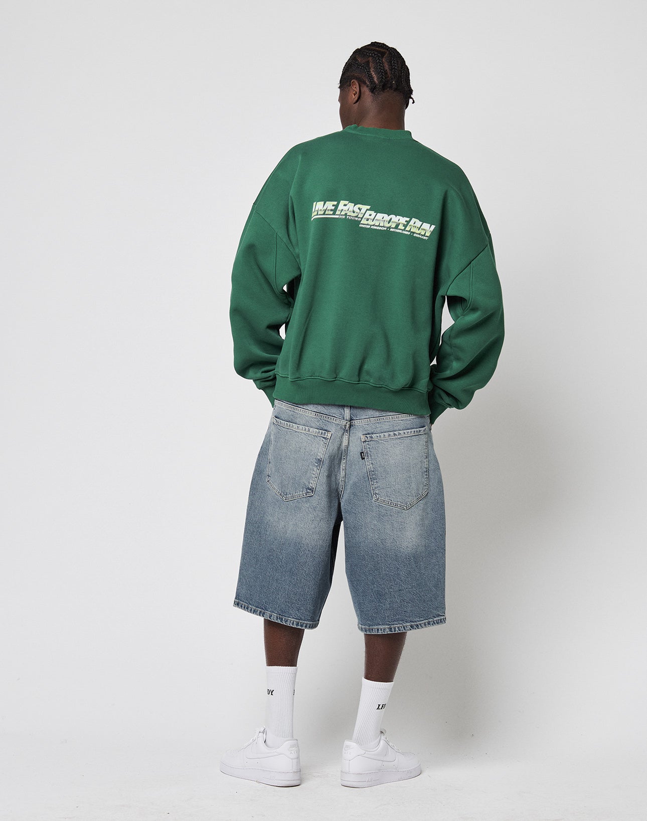 A person faces away, wearing the green LFDY Europe Run Sweater by LIVE FAST DIE YOUNG with back print, light blue knee-length denim shorts, white crew socks, and white sneakers, set against a plain white background.