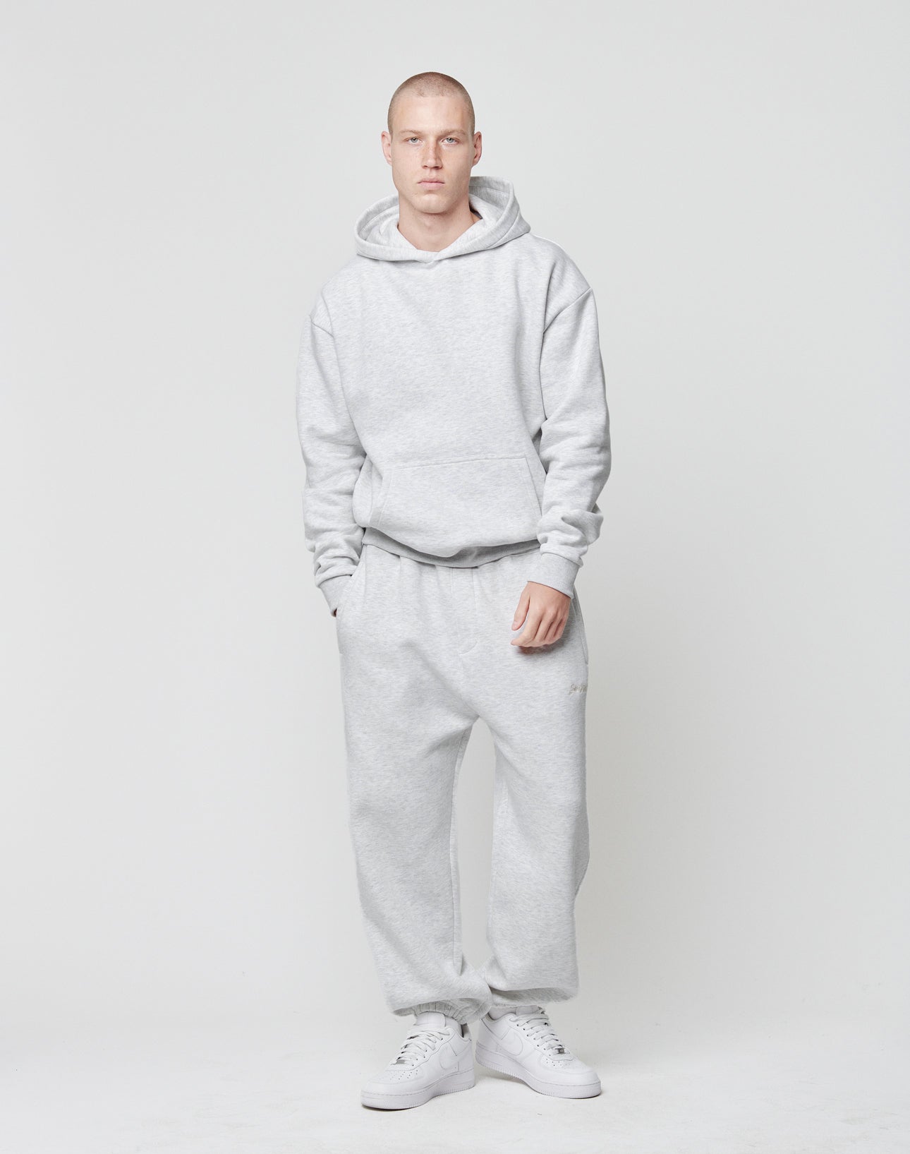 A person stands against a plain white background, wearing the LIVE FAST DIE YOUNG Basic Blank Hooded in an oversize fit light gray polyester-cotton blend, matching sweatpants, and white sneakers, hands in pockets and a neutral expression.