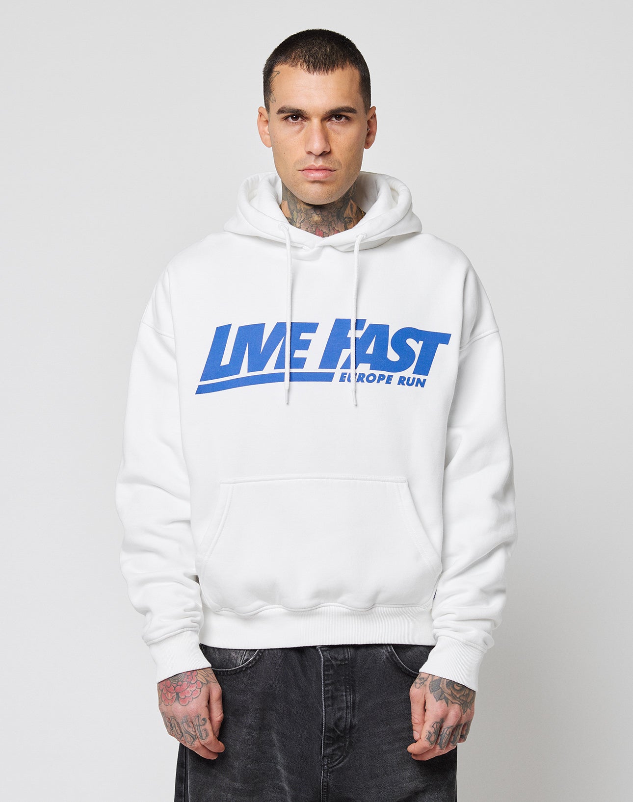 A person with short hair and visible tattoos wears the LIVE FAST DIE YOUNG Live Fast Europe Run Hooded, a white oversize hoodie with blue text, Kapuze, standing facing forward against a plain light background.