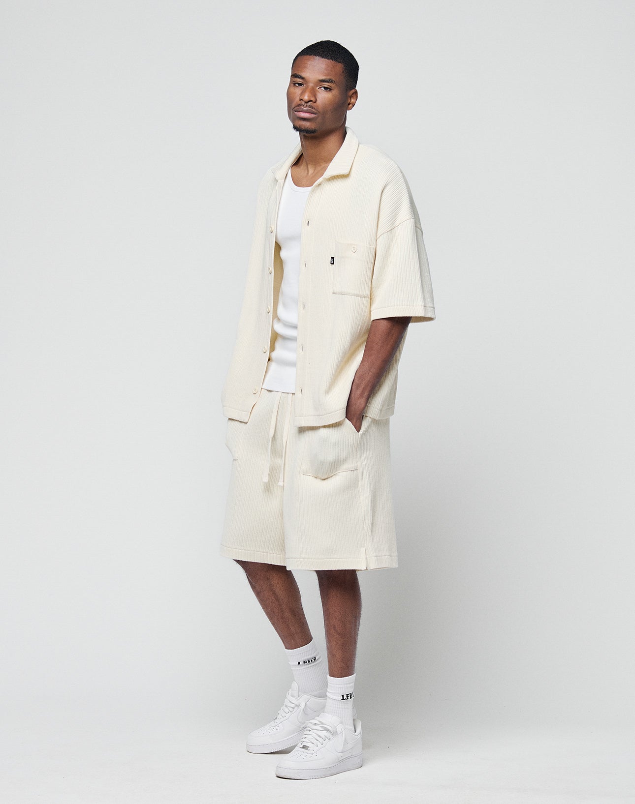 A man with a neutral expression wears LIVE FAST DIE YOUNG Basic Knit Shorts with elastic waistband, a cream shirt with patch pockets, white tank top, socks, and sneakers against a plain white background. He stands relaxed.