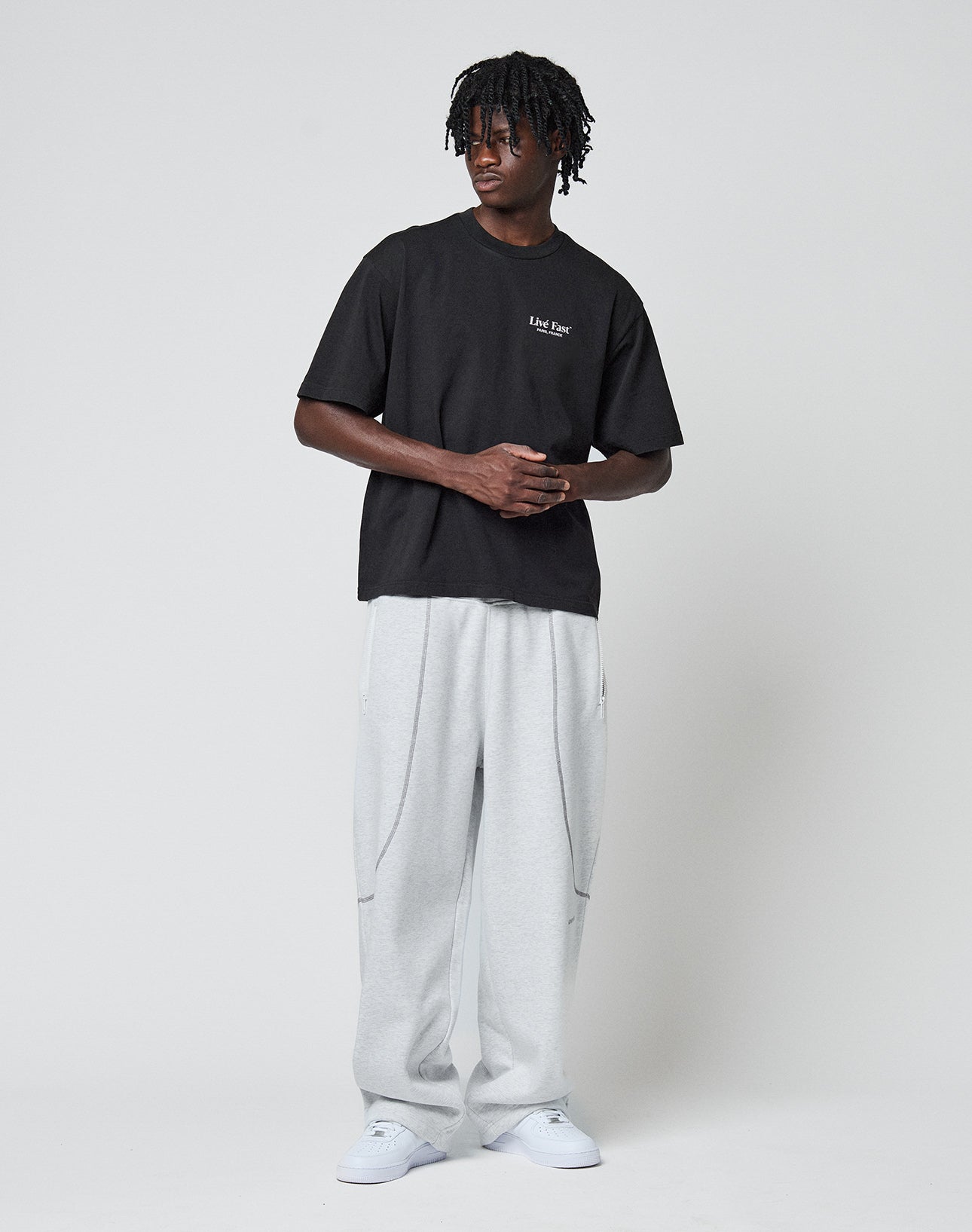 A man with short dreadlocks stands against a plain background, wearing the LIVE FAST DIE YOUNG LFDY Paris Tee—an oversize black t-shirt with small white text front and back—paired with light gray sweatpants and white sneakers.