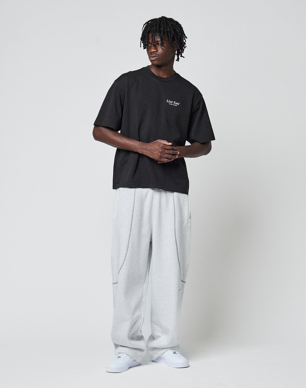 A man with short dreadlocks stands against a plain background, wearing the LIVE FAST DIE YOUNG LFDY Paris Tee—an oversize black t-shirt with small white text front and back—paired with light gray sweatpants and white sneakers.