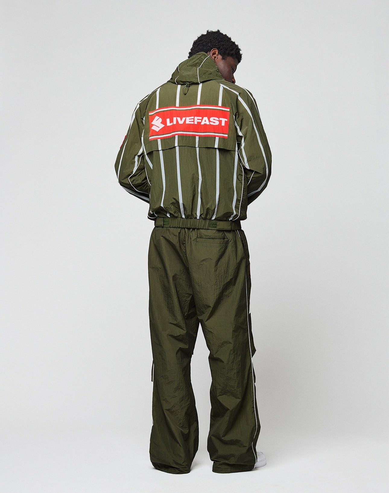 A person wears the LFDY Nylon Trackjacket by LIVE FAST DIE YOUNG—an olive green, oversize-fit jacket with white stripes, a stylized running figure, and a bold red LIVEFAST backprint for standout sporty style.