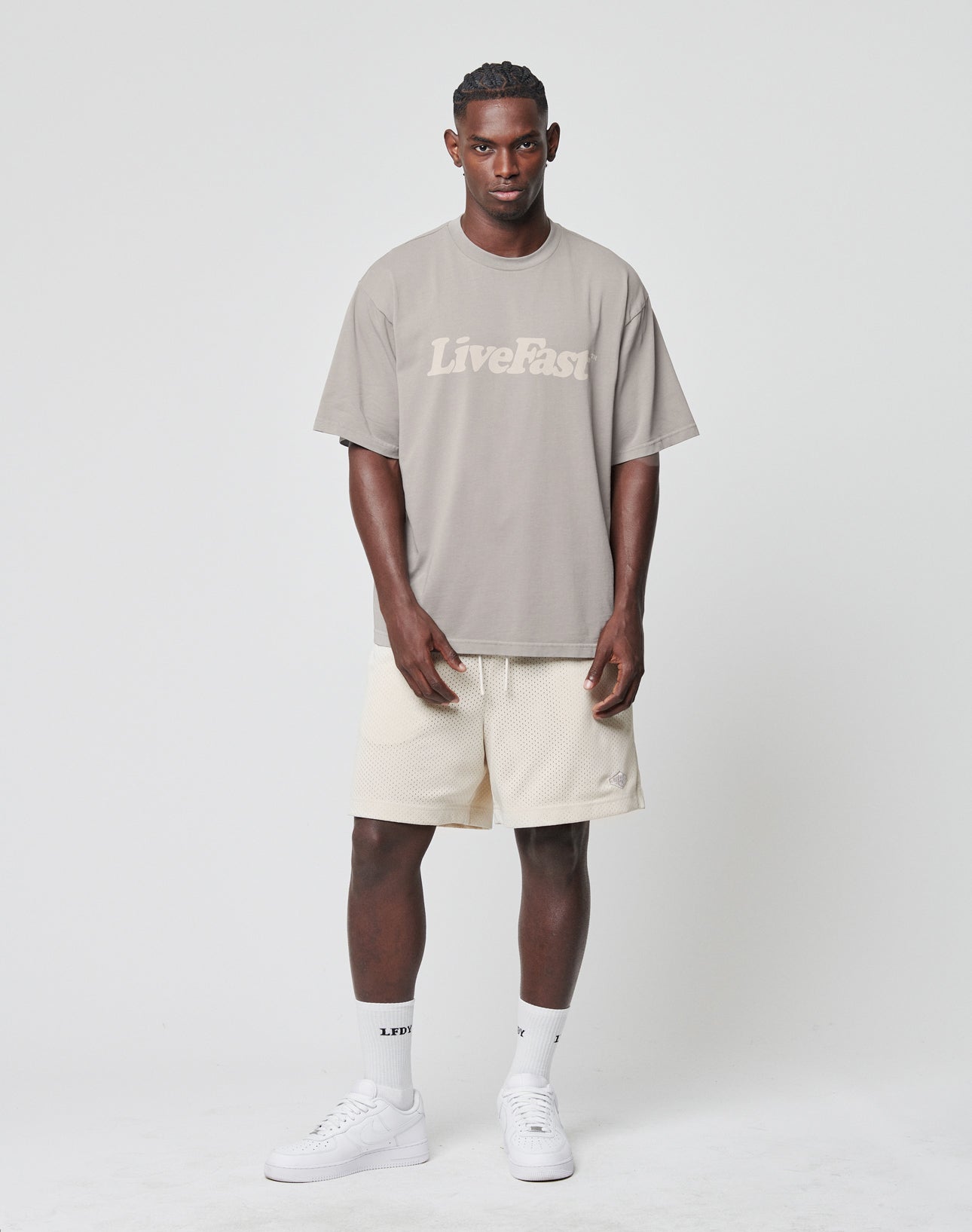 A man stands in a studio wearing a taupe LIVE FAST DIE YOUNG Basic Tee in vintage wash and oversize fit, light beige shorts, white LFE text socks, and white sneakers against a plain white background.