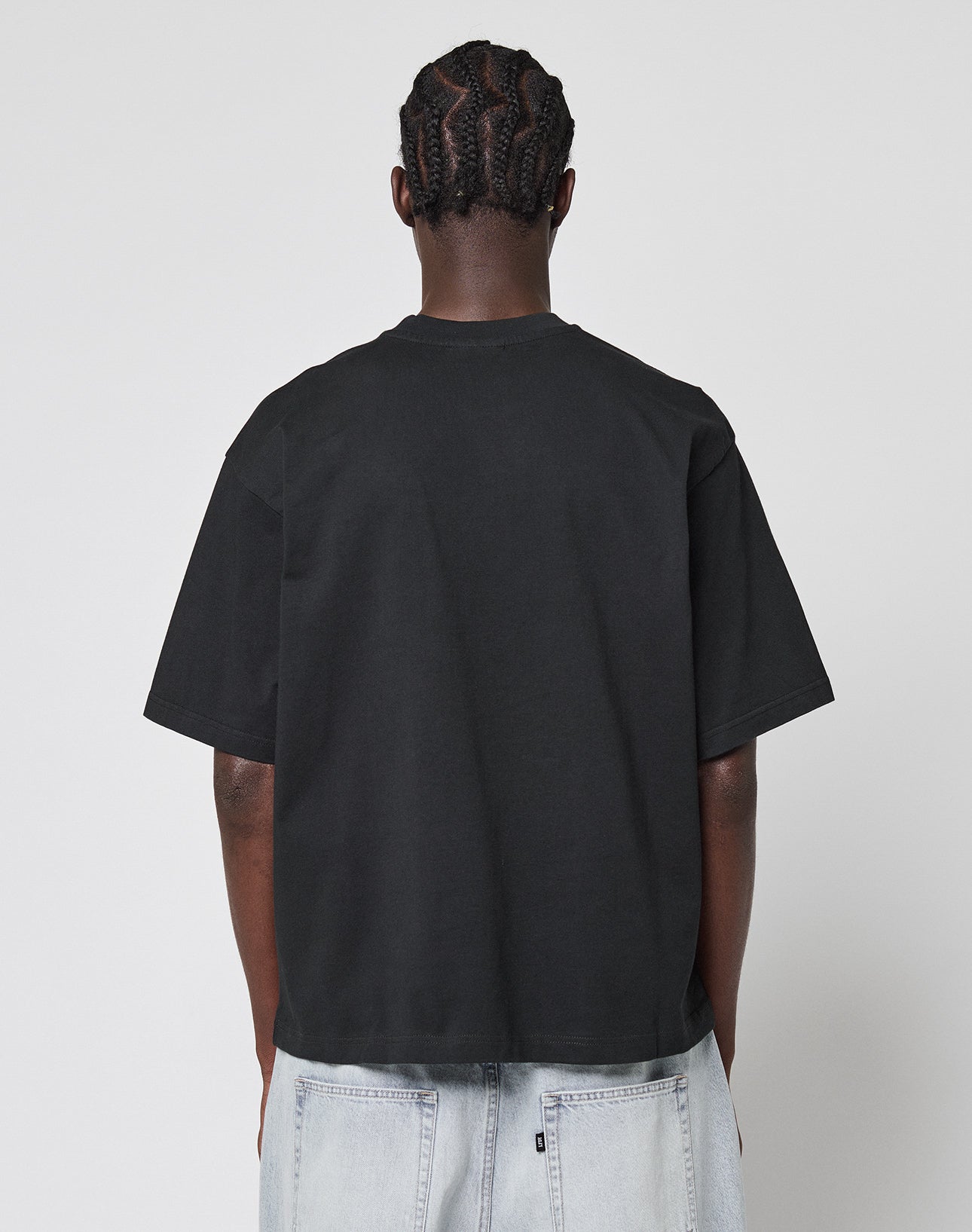 A person with braided hair stands with their back to the camera, wearing a LIVE FAST DIE YOUNG LFDY Triple Racing Tee in black and light-wash denim jeans against a plain white background.