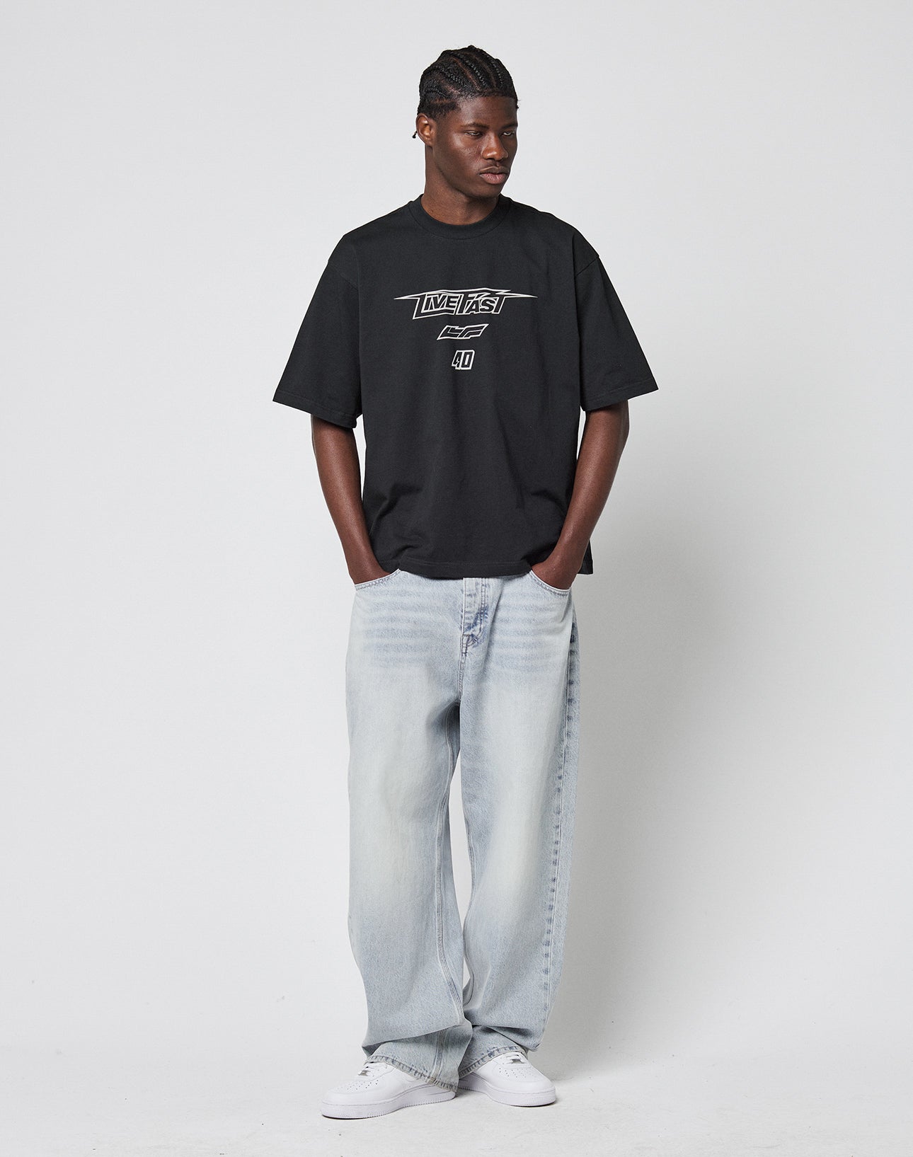 A person stands against a white background, wearing the LIVE FAST DIE YOUNG LFDY Triple Racing Tee—an oversize black graphic T-shirt made of 100% cotton—paired with light blue baggy jeans and white sneakers, hands in pockets, looking aside.