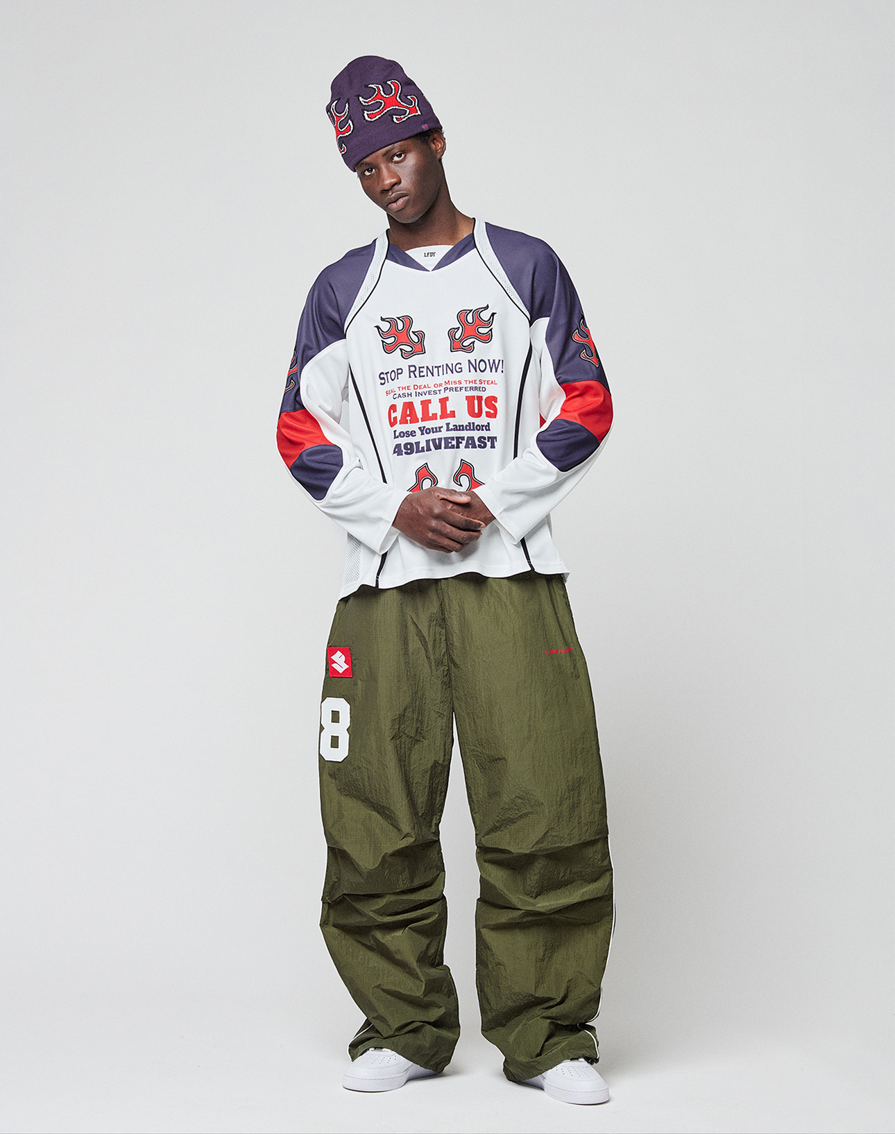A person stands against a plain backdrop wearing an oversize beanie, LIVE FAST DIE YOUNG green pants, white sneakers, and the LFDY Call Us Jersey long-sleeve graphic shirt. Their hands are in their pockets and their expression is neutral.