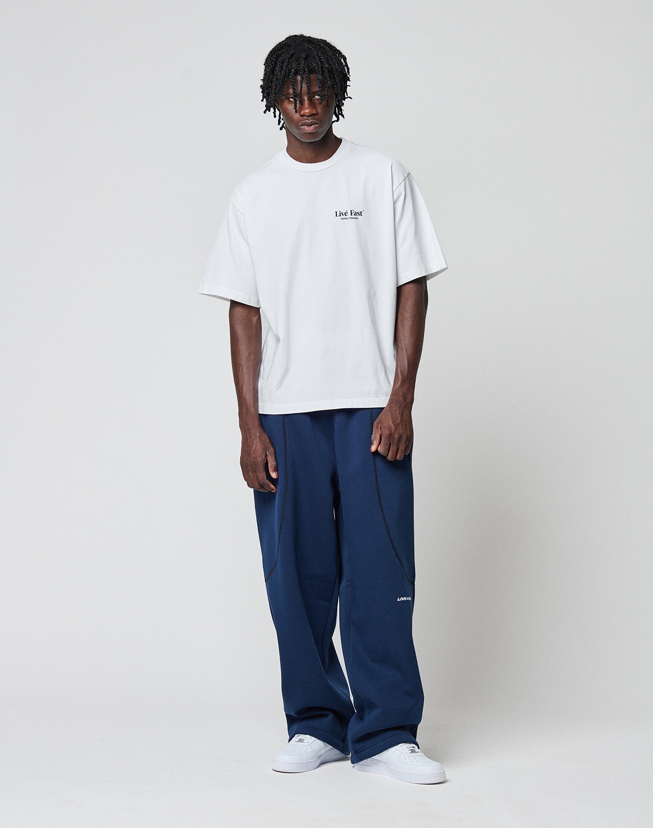 A man with short dreadlocks and a neutral expression stands against a plain background, wearing the LIVE FAST DIE YOUNG LFDY Paris Tee in white, navy wide-leg pants, and white sneakers. His arms are relaxed by his sides.