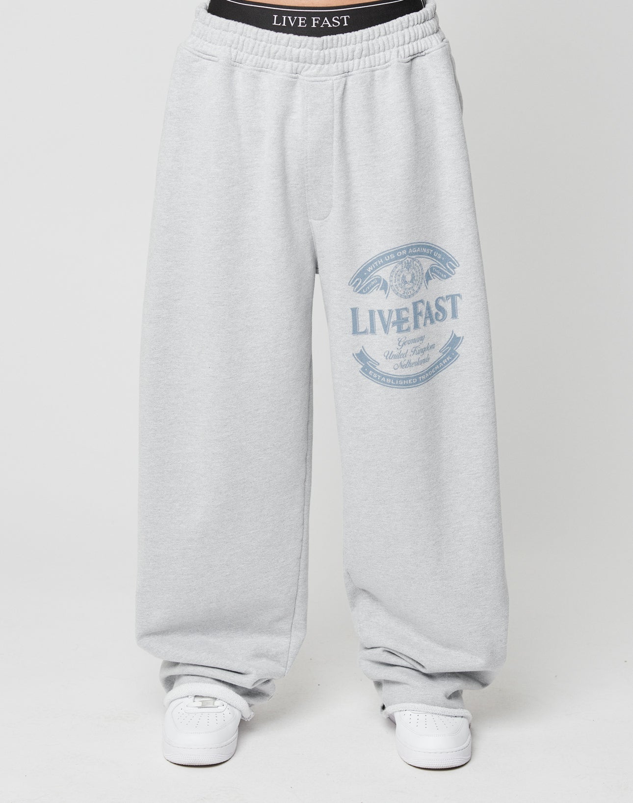 A person wears the Loyal Wide Jogger by LIVE FAST DIE YOUNG in light gray with a blue LIVE FAST graphic on the wide leg and “LIVE FAST” on the black elastic waistband, styled with white sneakers against a plain white background.