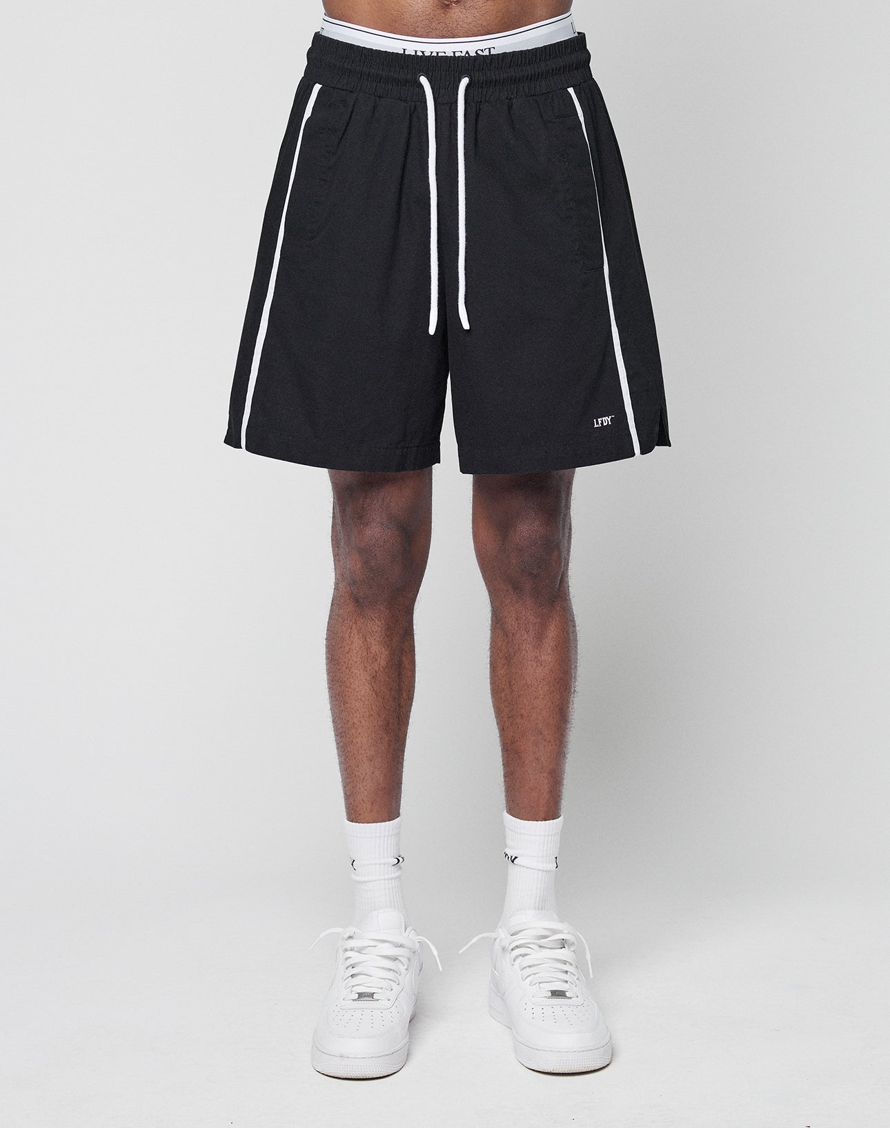 A person models LIVE FAST DIE YOUNG Basic Cotton Shorts—black with white stripes, elastic waistband, white drawstring, and logo print. White socks and sneakers complete the regular-fit outfit. Only the lower half is visible.