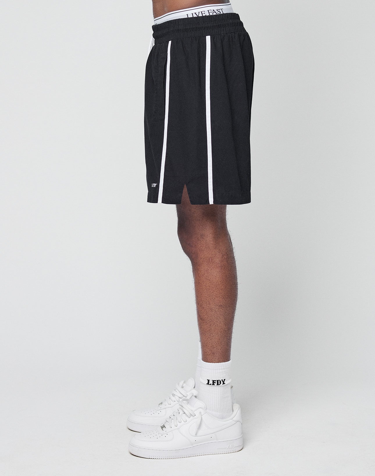 A person in profile wears LIVE FAST DIE YOUNG Basic Cotton Shorts with white side stripes and a logo print, paired with white crew socks featuring LFDY text and white Nike sneakers against a plain light background.