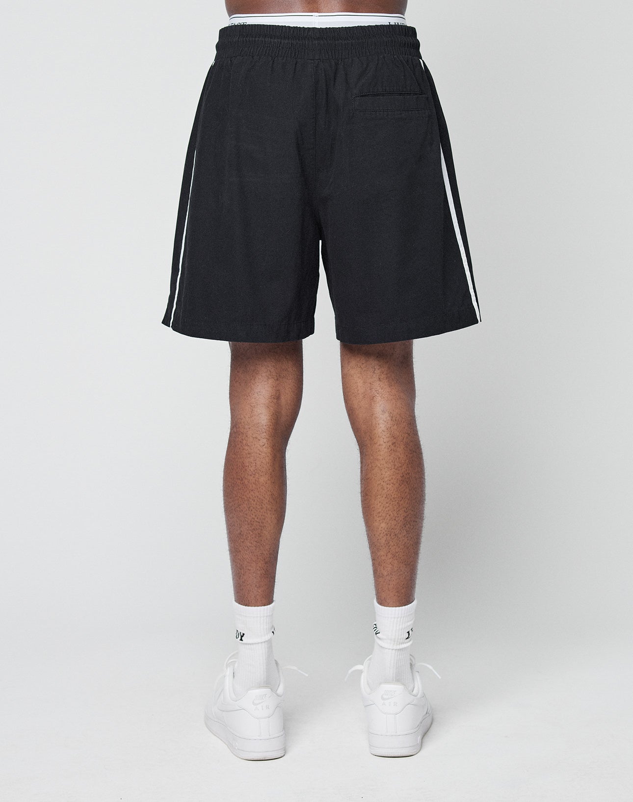 A person faces away, wearing LIVE FAST DIE YOUNG Basic Cotton Shorts in black with white side stripes and elastic waistband, paired with white socks and sneakers against a plain white background.