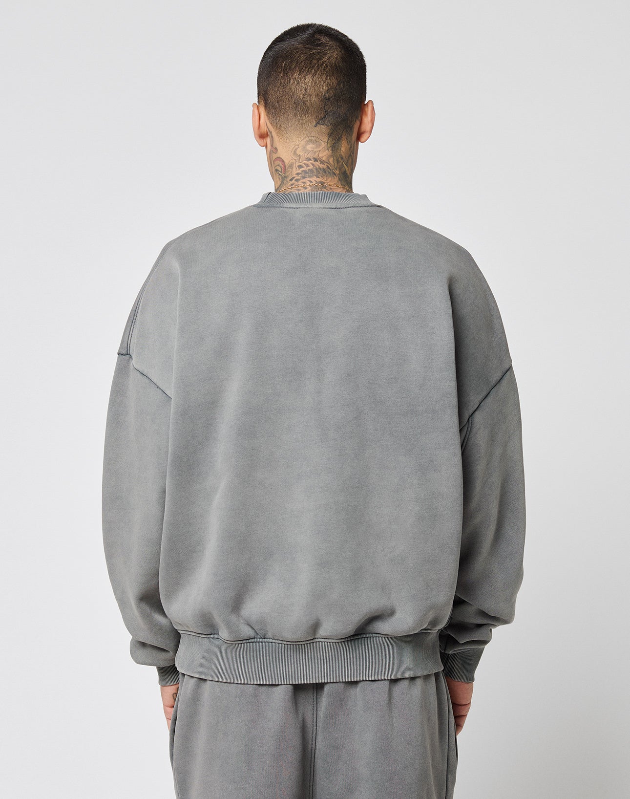 A person with short hair and visible neck tattoos faces away from the camera, wearing the LIVE FAST DIE YOUNG Live Fast Europe Run Sweater in faded gray with matching pants, set against a plain light background.