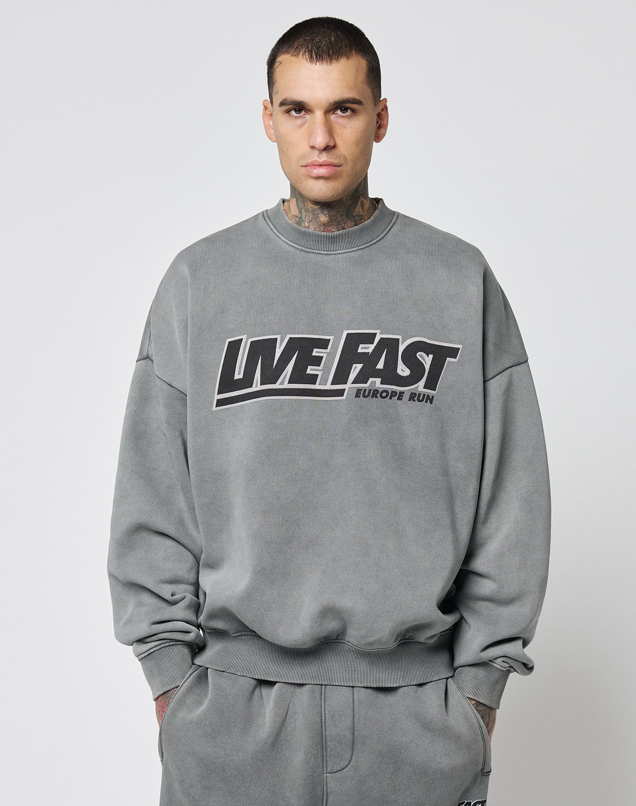 A man with short hair and neck tattoos models the LIVE FAST DIE YOUNG Live Fast Europe Run Sweater, a gray oversized sweatshirt with a bold front print, against a plain light background.