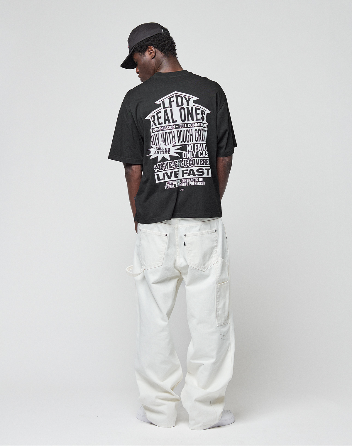 A person faces away, wearing a black LIVE FAST DIE YOUNG LFDY Real Ones Tee made of 100% Baumwolle with bold white front and back prints, a black cap, and loose-fitting white pants, set against a plain light grey background.