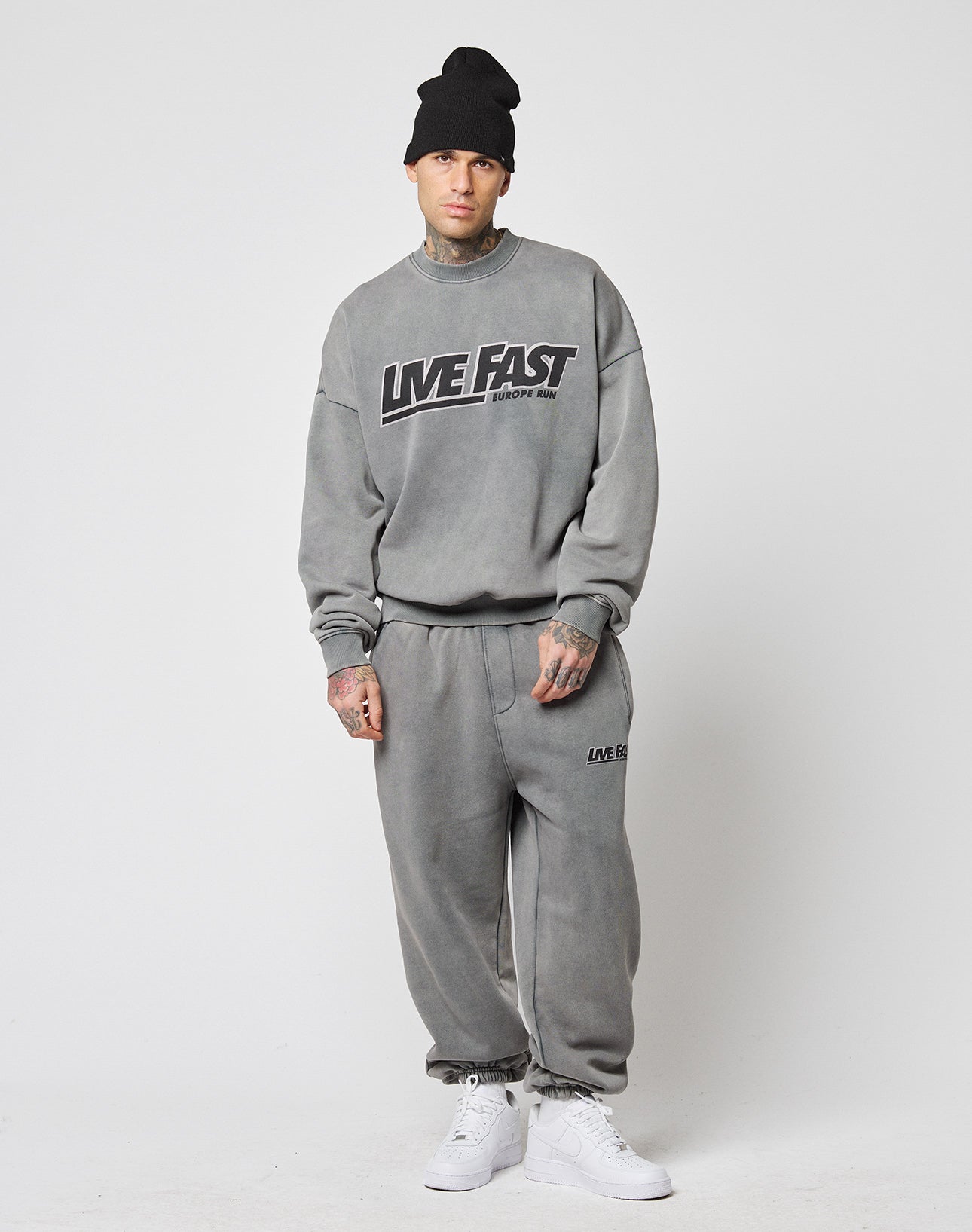 A man wearing a black beanie, LIVE FAST DIE YOUNGs grey Live Fast Europe Run Sweater in an oversize fit with front print, matching grey sweatpants, and white sneakers stands against a plain white background. Tattoos appear on his neck and hands.