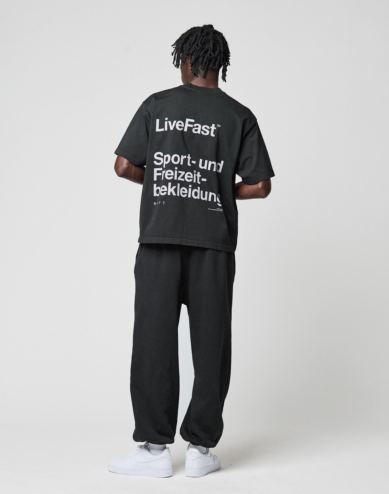 A person faces away wearing the LIVE FAST DIE YOUNG Basic Trademark Tee in black, featuring front and back white LiveFast text, paired with matching black pants and white sneakers, set against a plain light background.