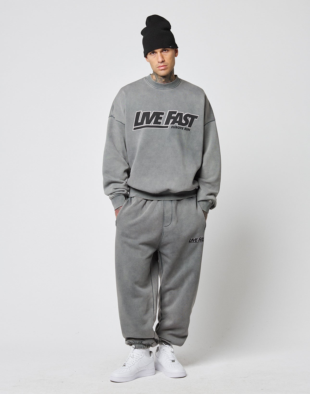 A person stands against a white background wearing a black beanie, grey sweatshirt, LIVE FAST DIE YOUNG LFDY Europe Run Jogger sweatpants with elastic cuffs, and white sneakers.