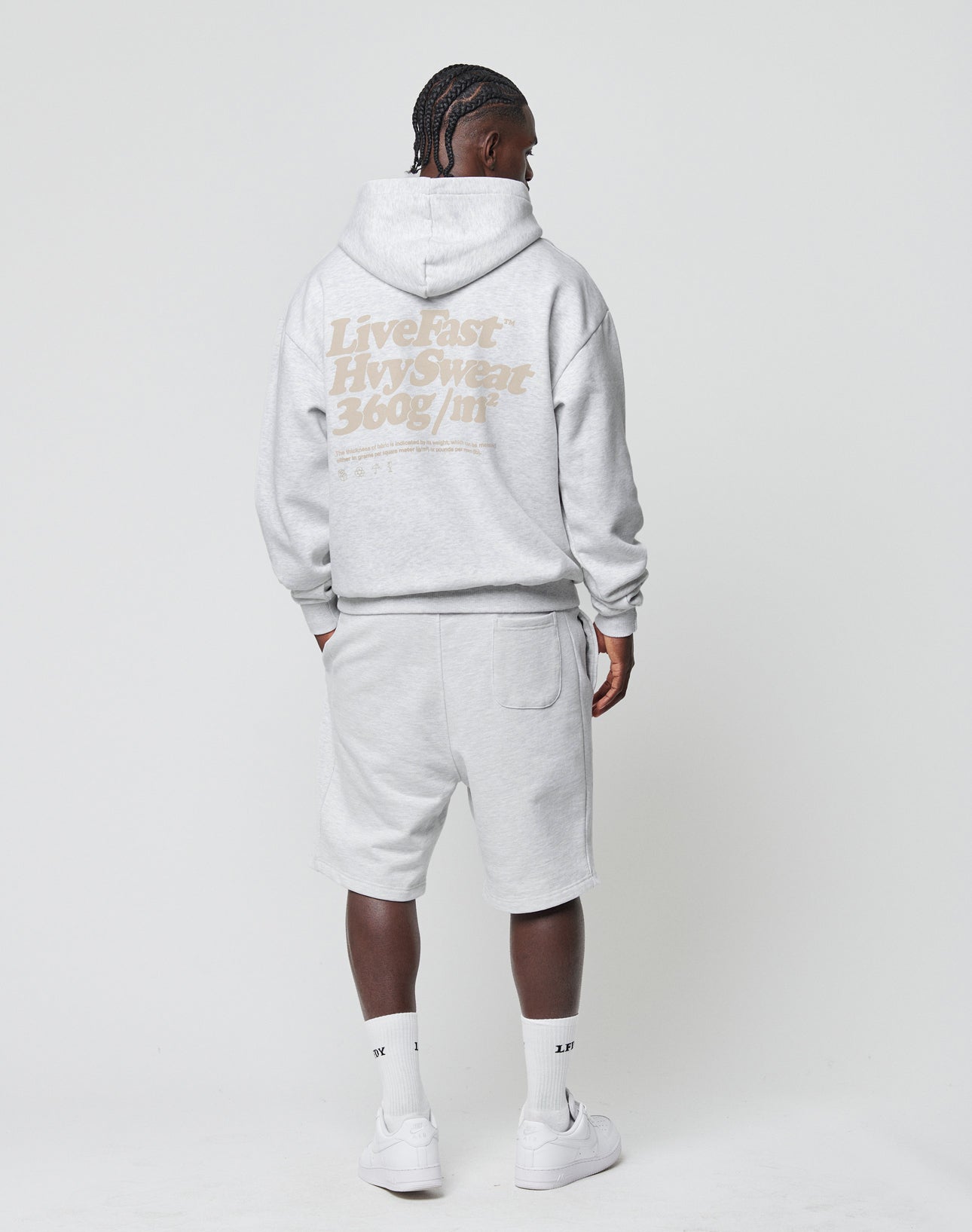 A person, seen from behind against a white background, wears the LIVE FAST DIE YOUNG Basic 360 Hooded in light gray Oversize Fit with matching shorts, white socks, and sneakers. HvySweat 360g/m² is printed on the back.