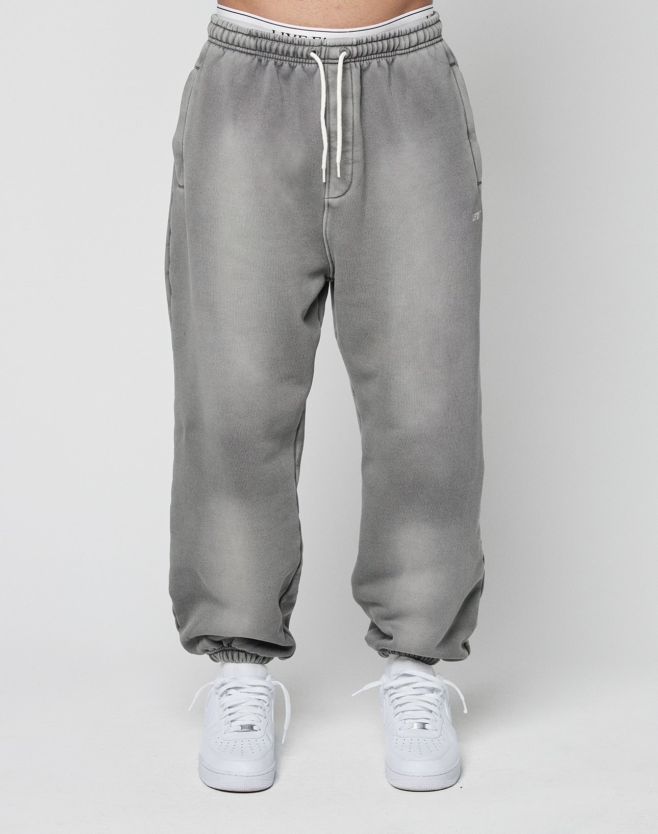 A person models the LIVE FAST DIE YOUNG Basic Loose Jogger—light gray with a subtle logo and elastic waistband—paired with white sneakers, standing against a plain light gray background.