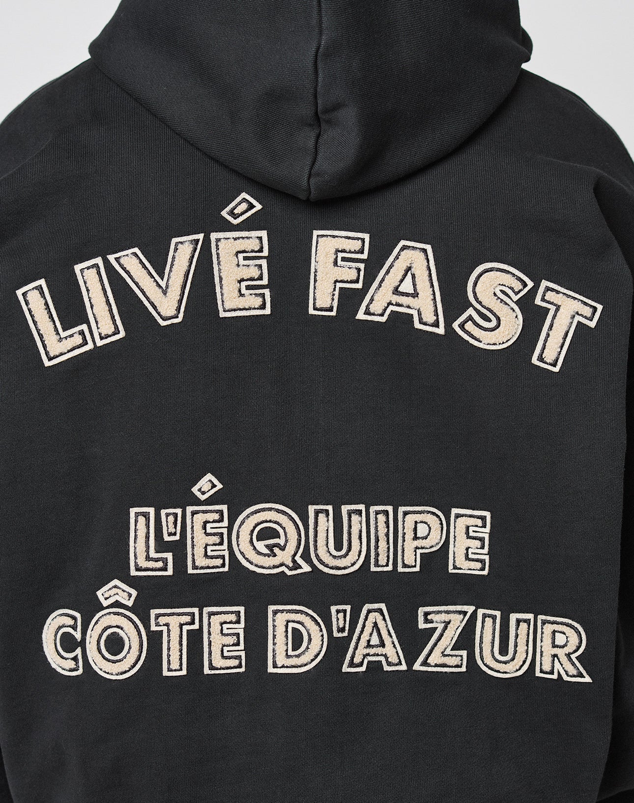 The LFDY LÉquipe Hooded Zip by LIVE FAST DIE YOUNG is a black oversized hoodie featuring beige chenille text on the back: LIVE FAST and LÉQUIPE CÔTE DAZUR in bold, outlined letters.