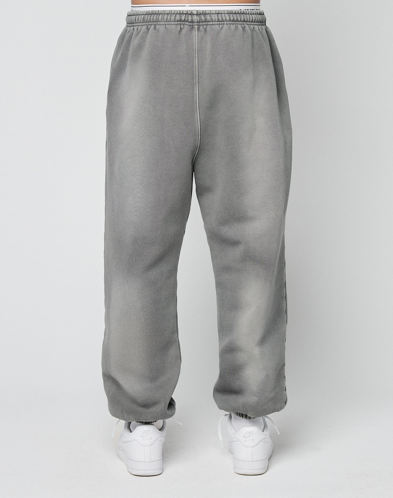 A person models the LIVE FAST DIE YOUNG Basic Loose Jogger—gray sweatpants with an elastic waistband—paired with white athletic shoes, standing against a plain light background.