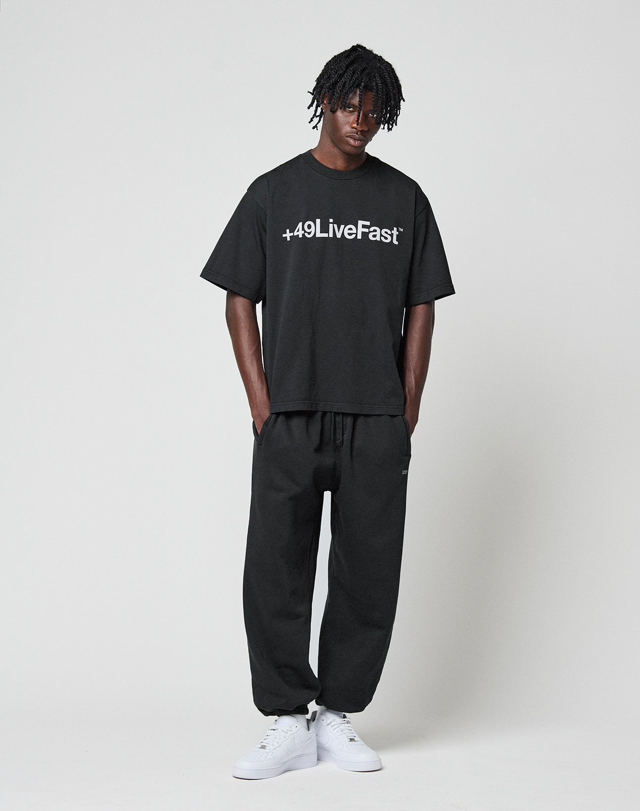 A person stands against a plain background wearing the LIVE FAST DIE YOUNG Basic +49 Tee, an oversize black 100% cotton shirt with +49LiveFast front print, paired with black joggers and white sneakers. They have short hair and relaxed hands-in-pockets posture.