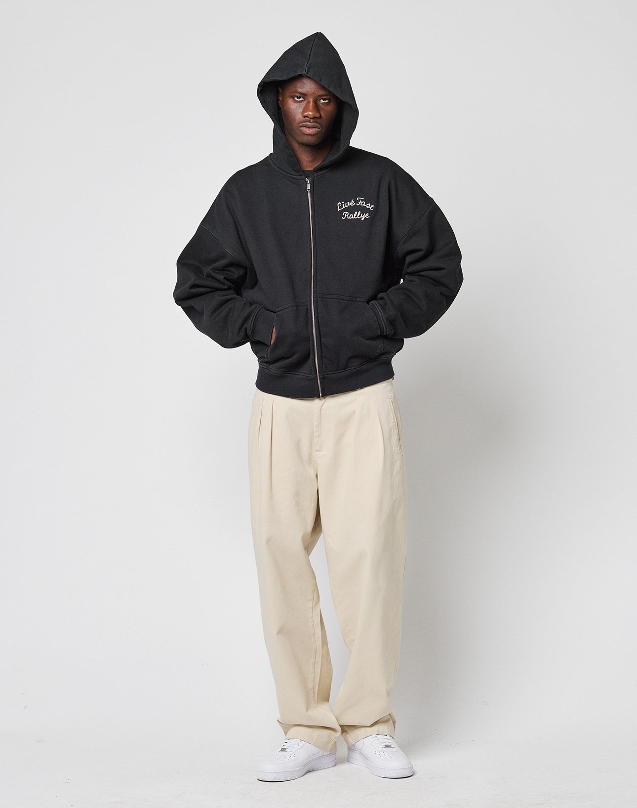 A person wearing the LIVE FAST DIE YOUNG LFDY LÉquipe Hooded Zip in black with the hood up, beige pleated pants, and white sneakers stands against a plain white background with hands in the hoodie pockets.