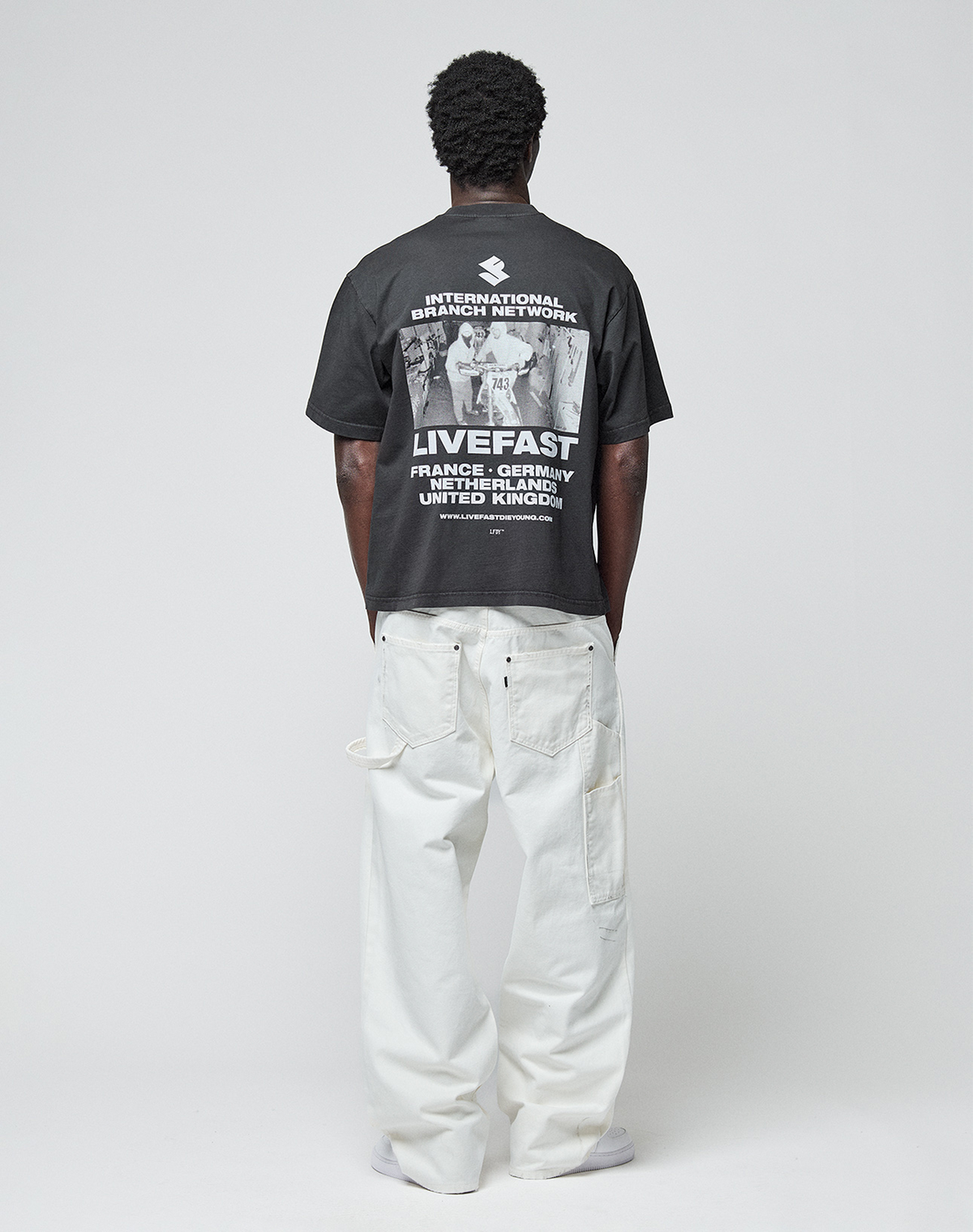 A person faces away, wearing the LIVE FAST DIE YOUNG LFDY Grand Theft Tee—an oversized black T-shirt with bold white graphics on front and back—paired with loose white pants against a plain light background.