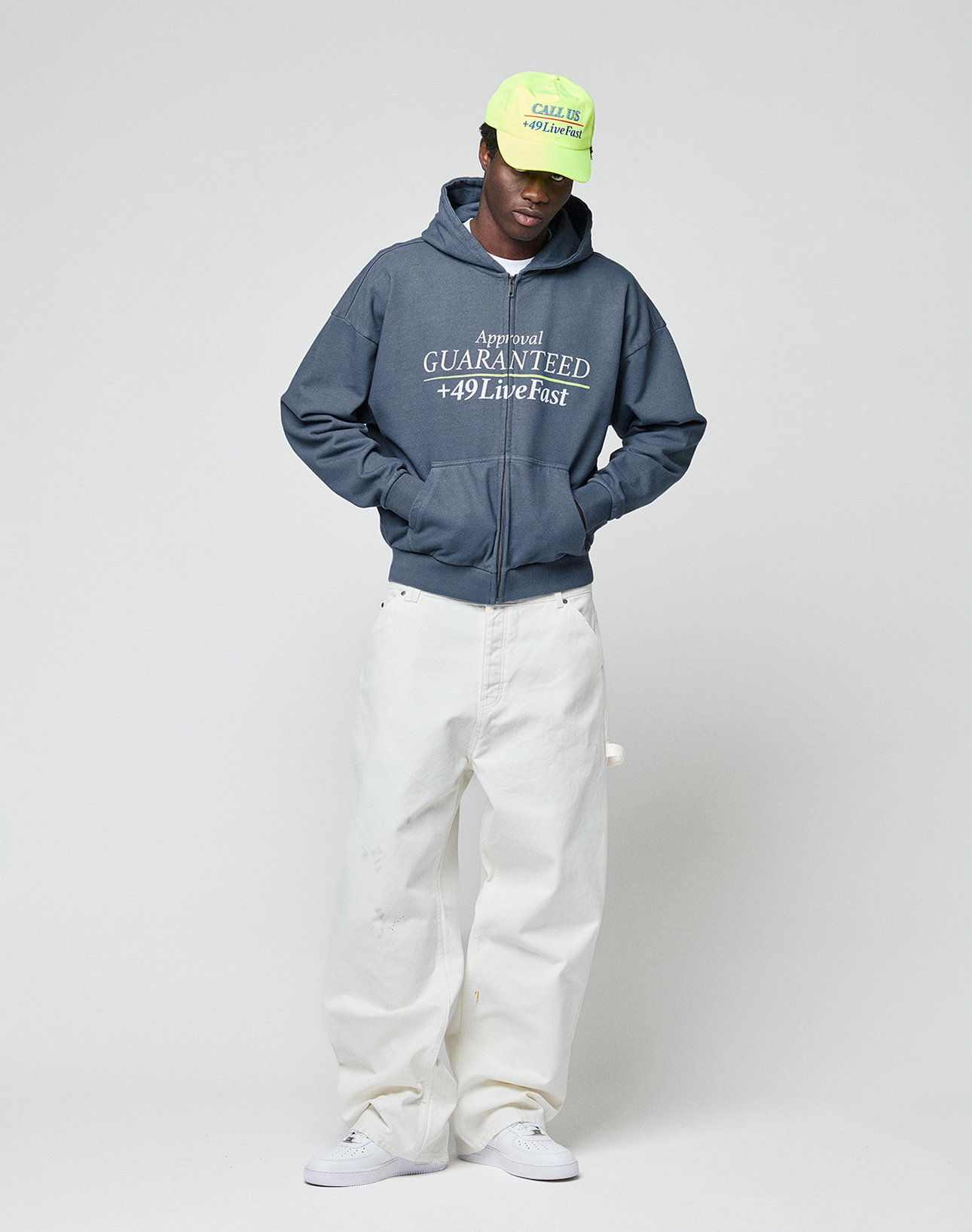 A person wearing a neon green cap, loose oversized white pants, white sneakers, and the LIVE FAST DIE YOUNG LFDY Guaranteed Hooded Zip with white frontprint stands against a plain light gray background, looking down with hands in pockets.