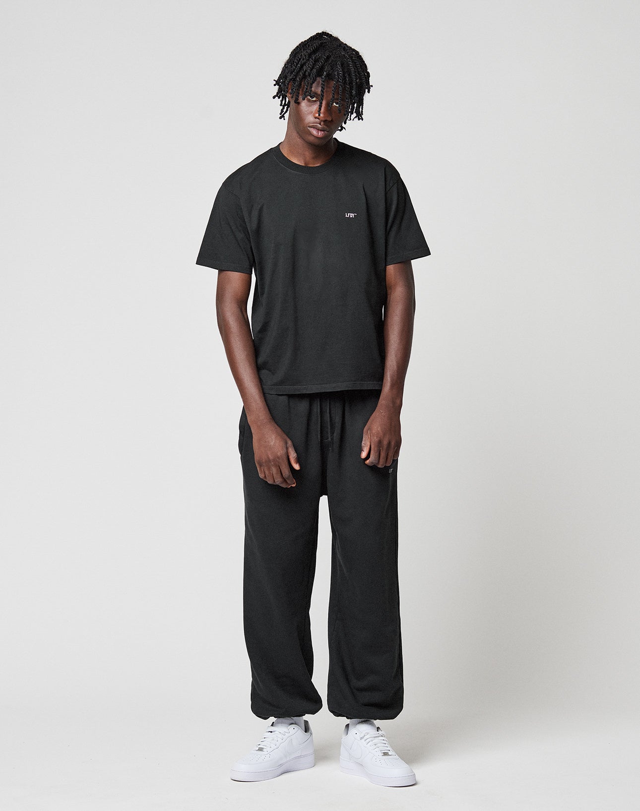 A man with short dreadlocks stands against a white background, wearing LIVE FAST DIE YOUNGs Basic Loose Jogger and a matching black t-shirt. He pairs the look with white sneakers, arms relaxed at his sides and a calm expression.