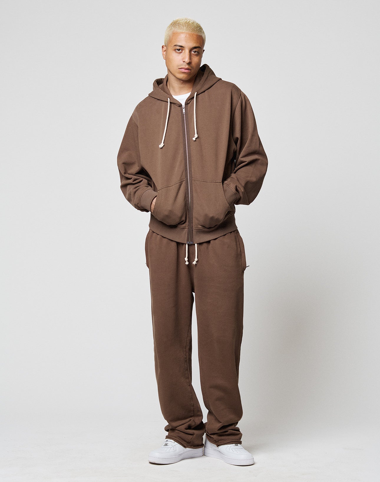A person with short blond hair wears the LFDY Lightweight Jogger by LIVE FAST DIE YOUNG—an oversize fit brown zip-up hoodie with white drawstring and matching brown French Terry sweatpants. They stand hands-in-pockets, wearing white sneakers against a plain background.