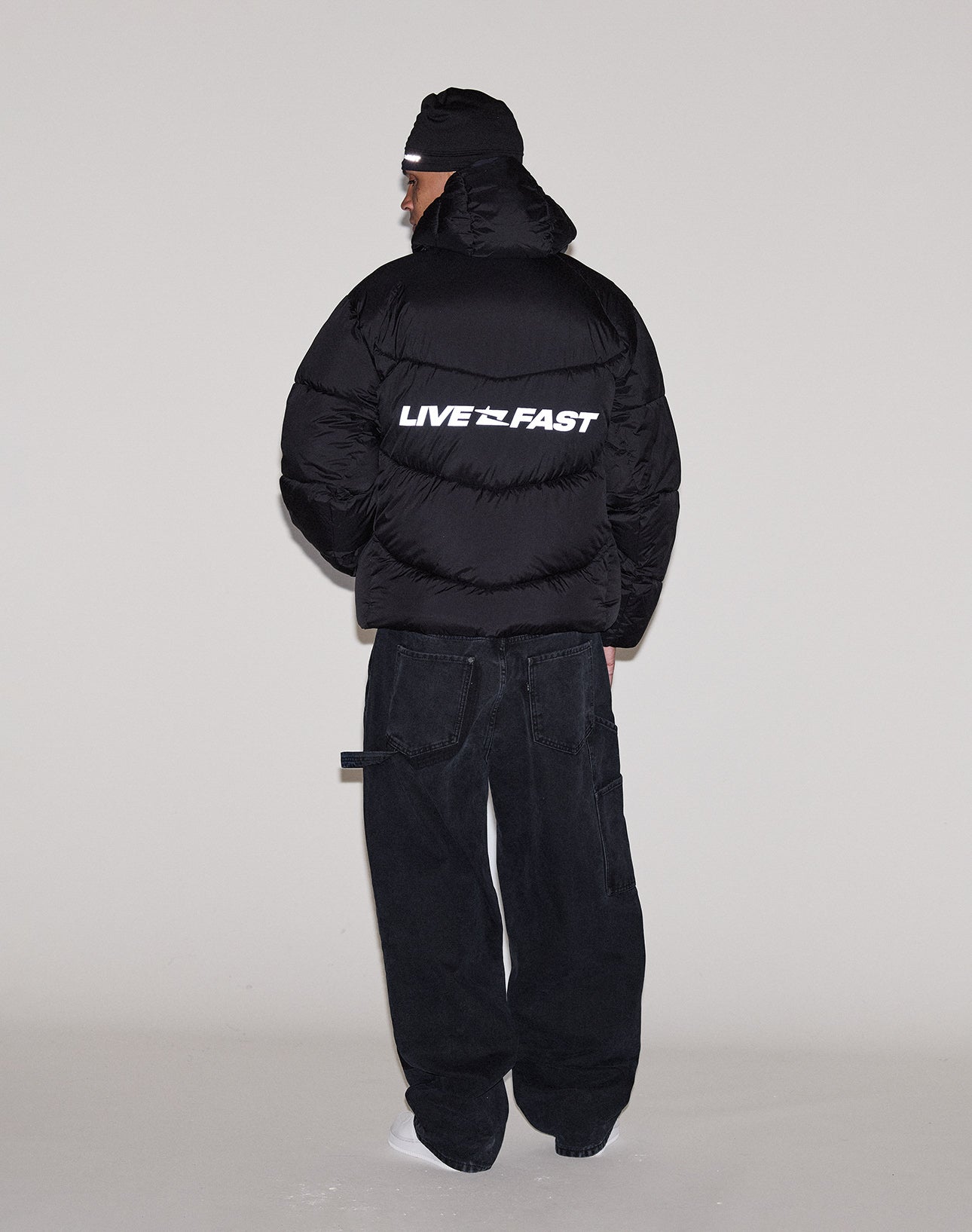 A person faces away, wearing the LIVE FAST DIE YOUNG Live Fast Puffer Jacket in black with a white reflective "LIVE FAST" print on the back, styled with black loose pants, a black beanie, and white shoes against a plain light background.