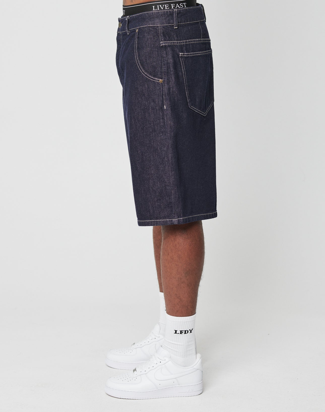 A person stands in profile wearing LFDY Jorts by LIVE FAST DIE YOUNG—dark denim, baggy knee-length Baumwolle shorts with LIVE FAST on the waistband and a Flag-Label detail—paired with LFDY logo white socks and white sneakers against a plain white background.