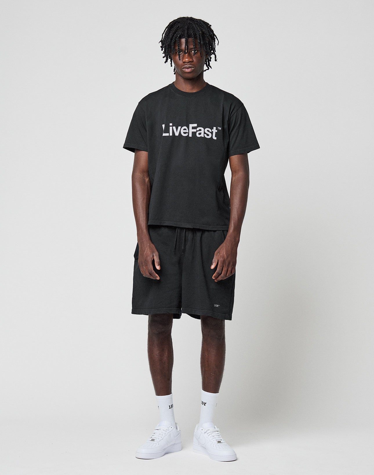 A man stands against a white background, wearing a black LIVE FAST DIE YOUNG logo t-shirt with an oversized fit, Basic Sweat Shorts, white socks, and white sneakers. He faces forward with a neutral expression and relaxed arms.